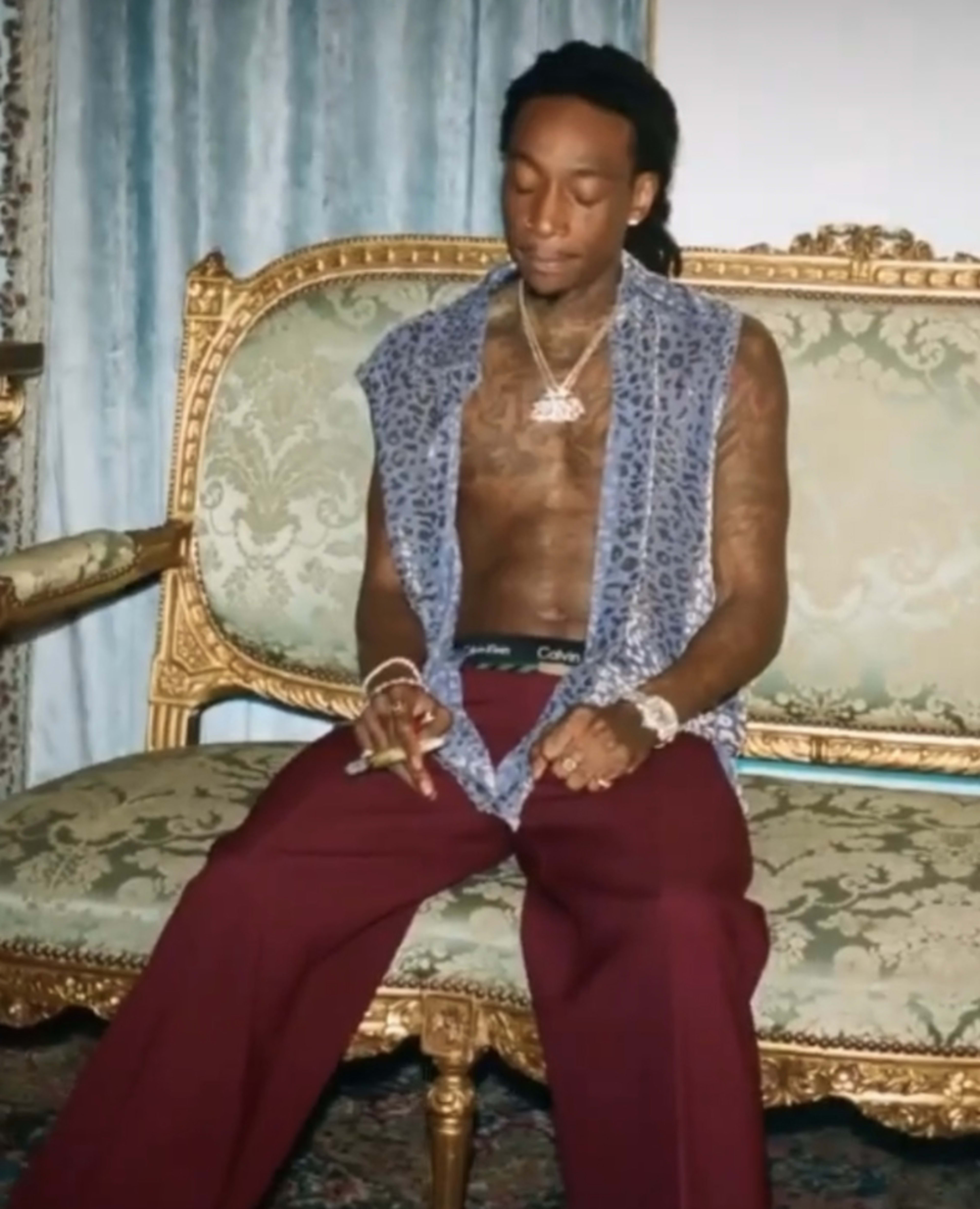 A man with tattoos sits on a vintage, ornate sofa, wearing loose-fitting pants and a patterned open shirt.