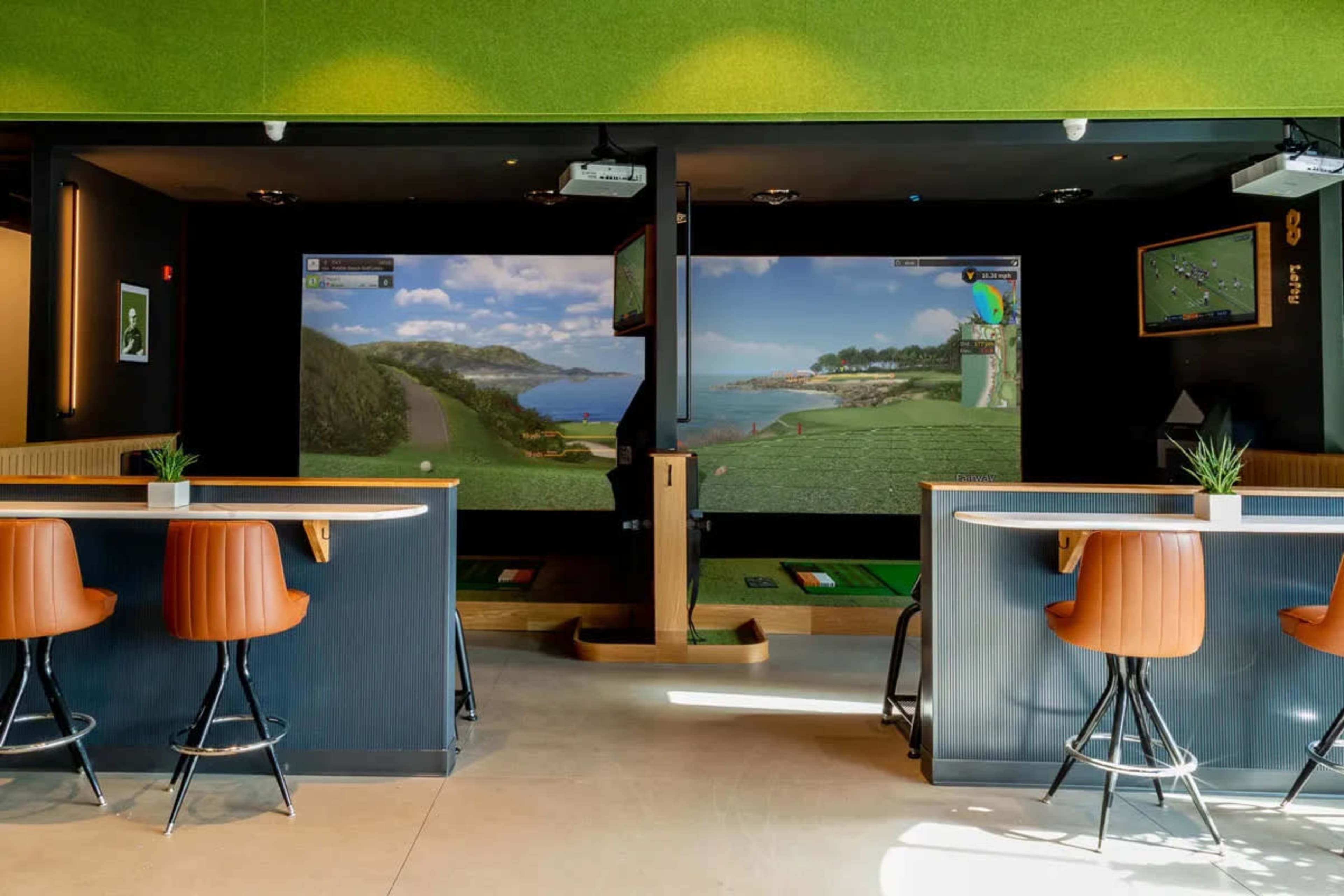 An indoor golf simulator space with two large screens displaying golf course scenes, along with golf mats and seating around a bar area.