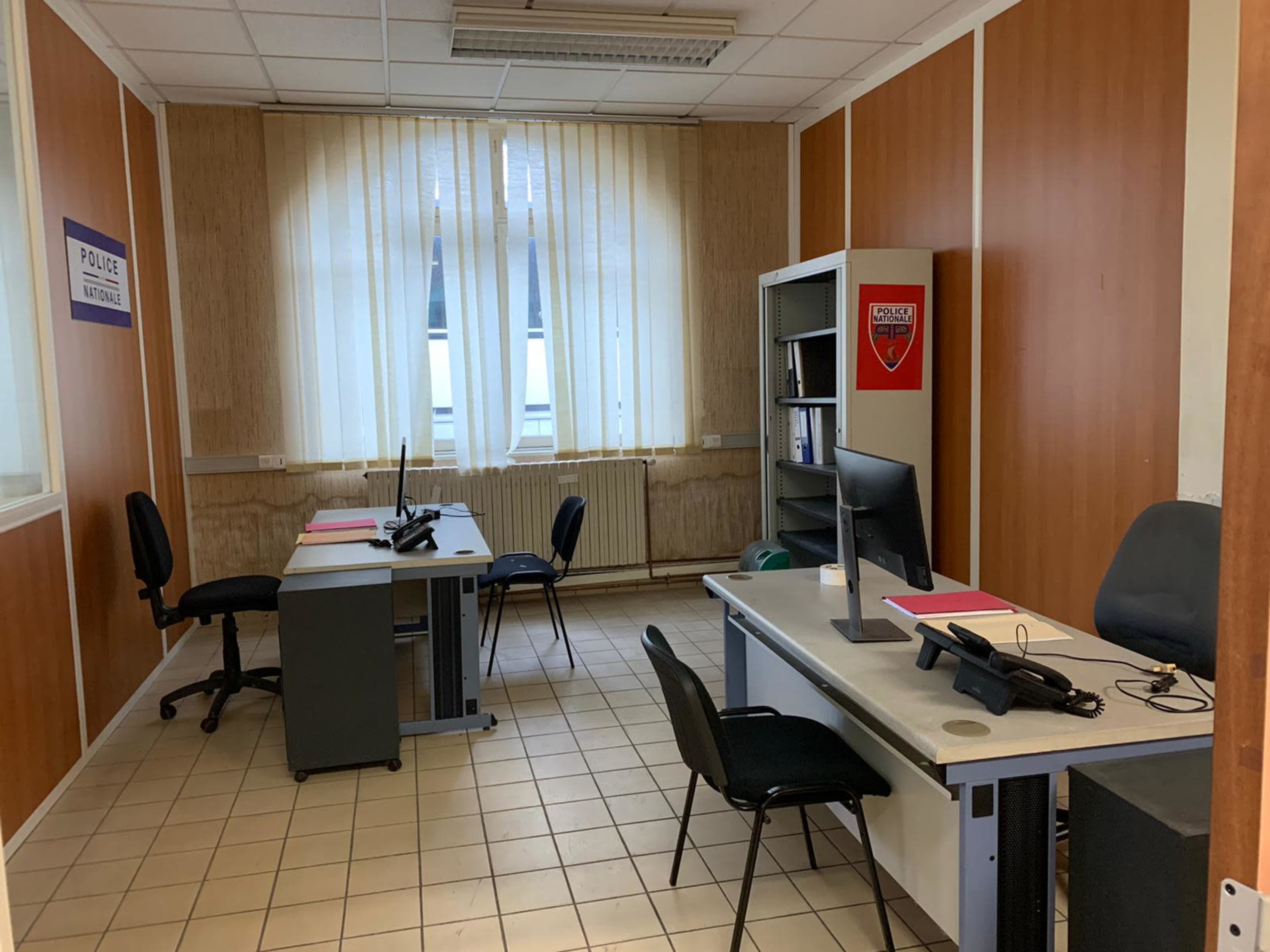 A sparsely furnished office with two desks, chairs, a filing cabinet, and large windows covered with light-colored curtains.