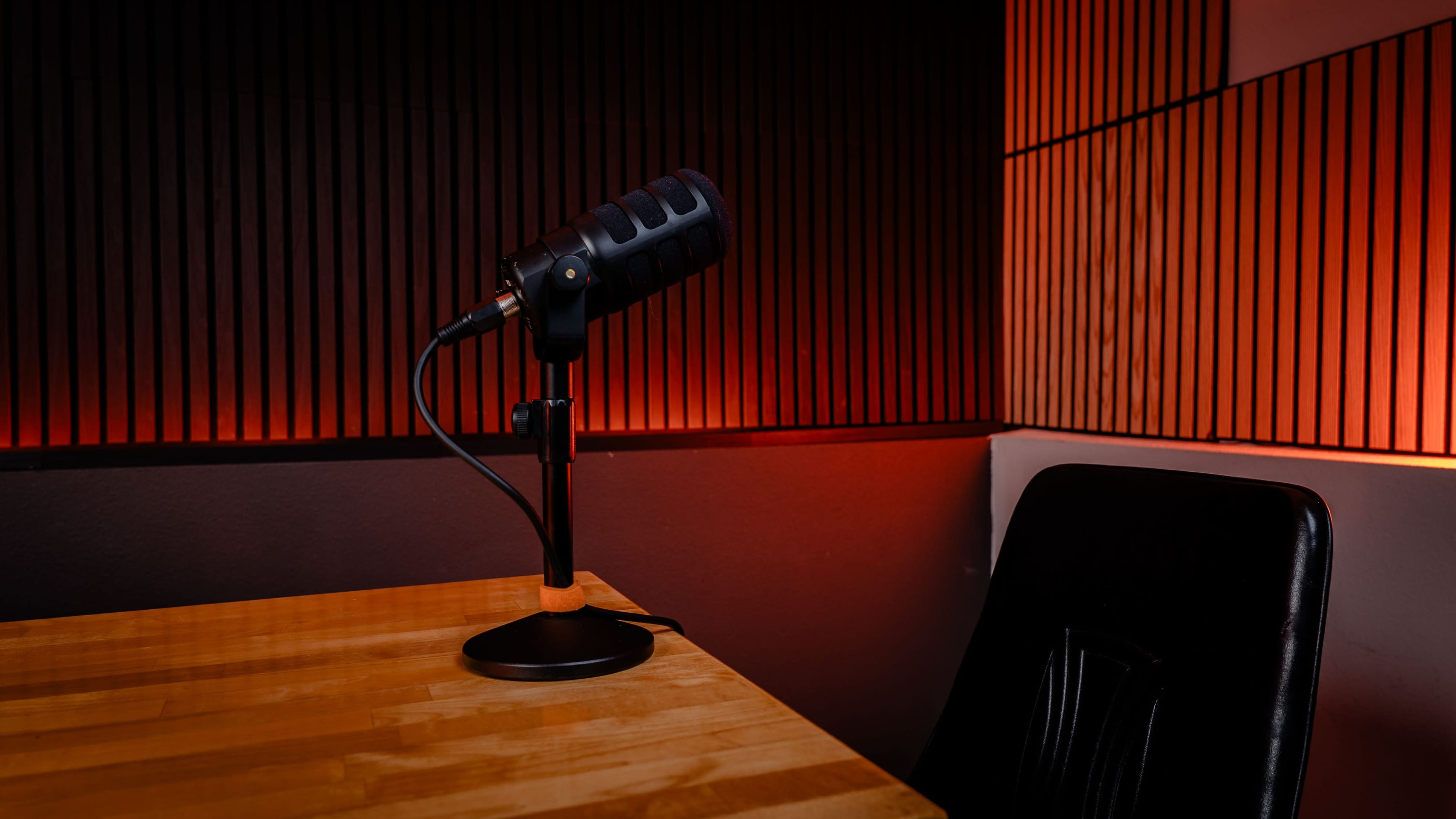 The image shows a black microphone on a sturdy stand positioned on a wooden table, with a black chair nearby and ambient orange lighting illuminating the backdrop of vertical wooden slats.