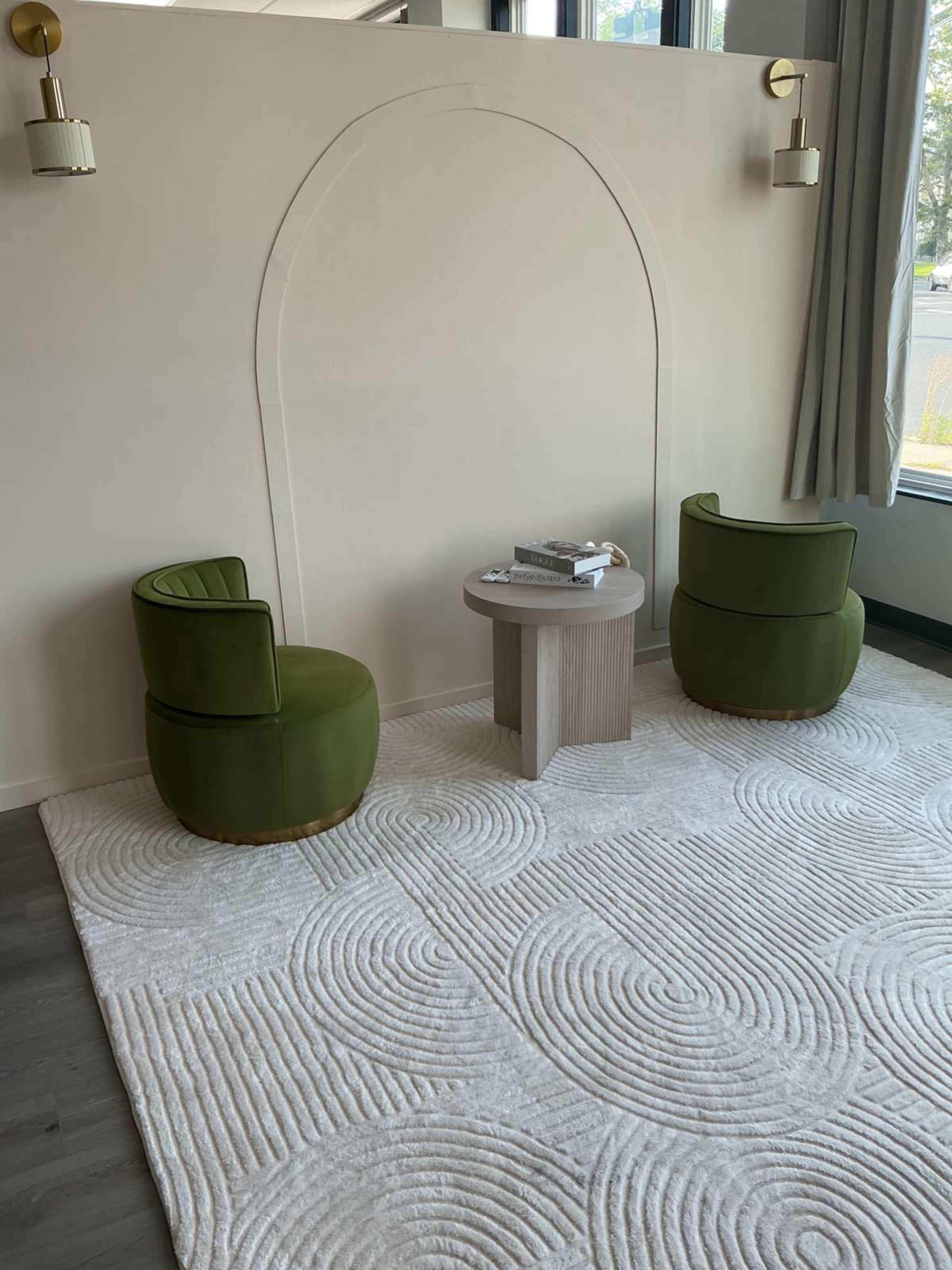 The image shows two green upholstered chairs positioned around a small round table on a textured cream-colored rug, against a light-colored wall with an arch design.
