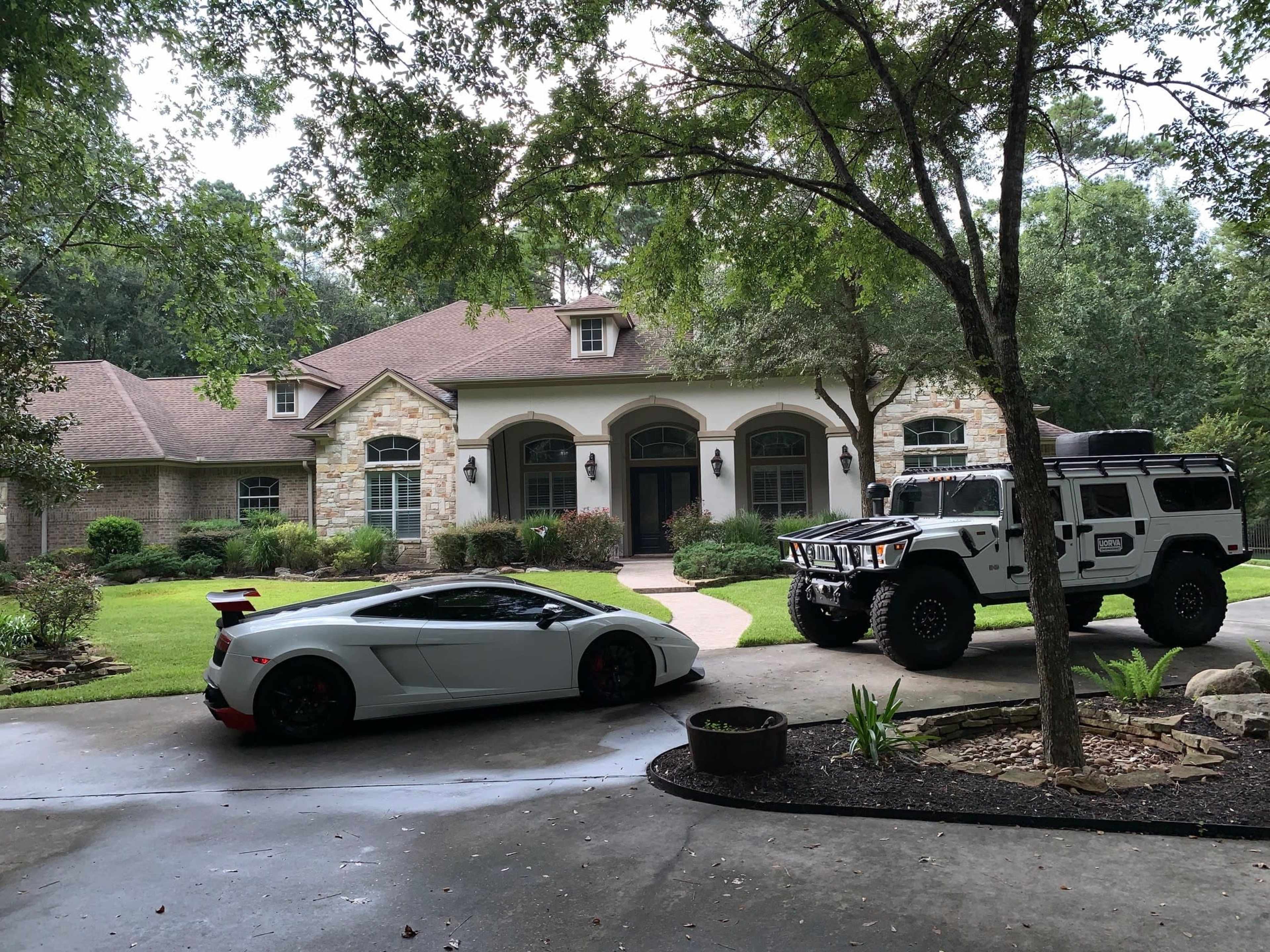 A white Lamborghini and a white Hummer are parked in front of a large, stone-accented residential house surrounded by greenery.