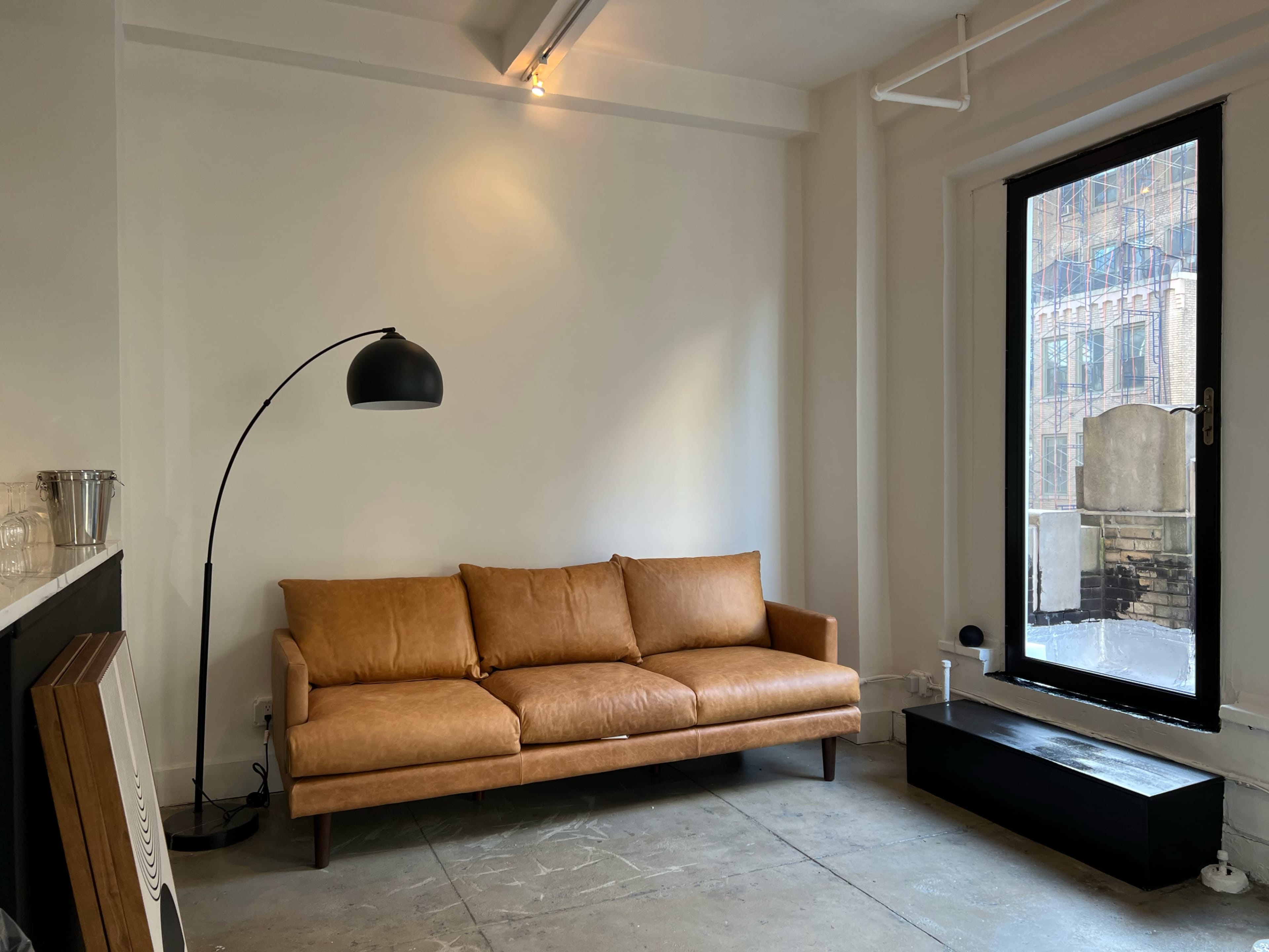 A tan leather sofa is positioned against a plain white wall next to a large black framed window.