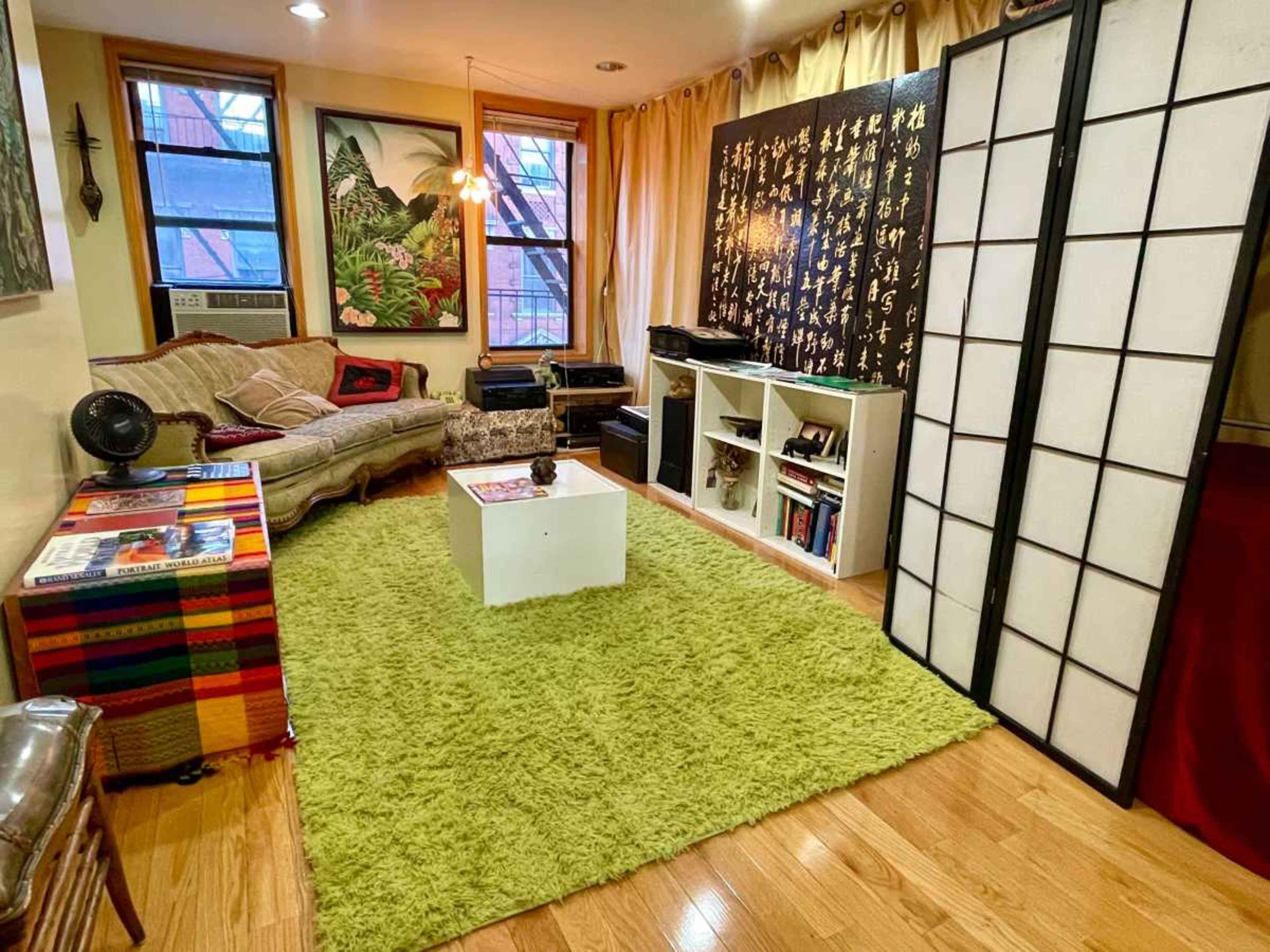 The image shows a cozy living room with a green rug, a sectional sofa, a coffee table, and a large bookshelf against a wall decorated with black and gold artwork.