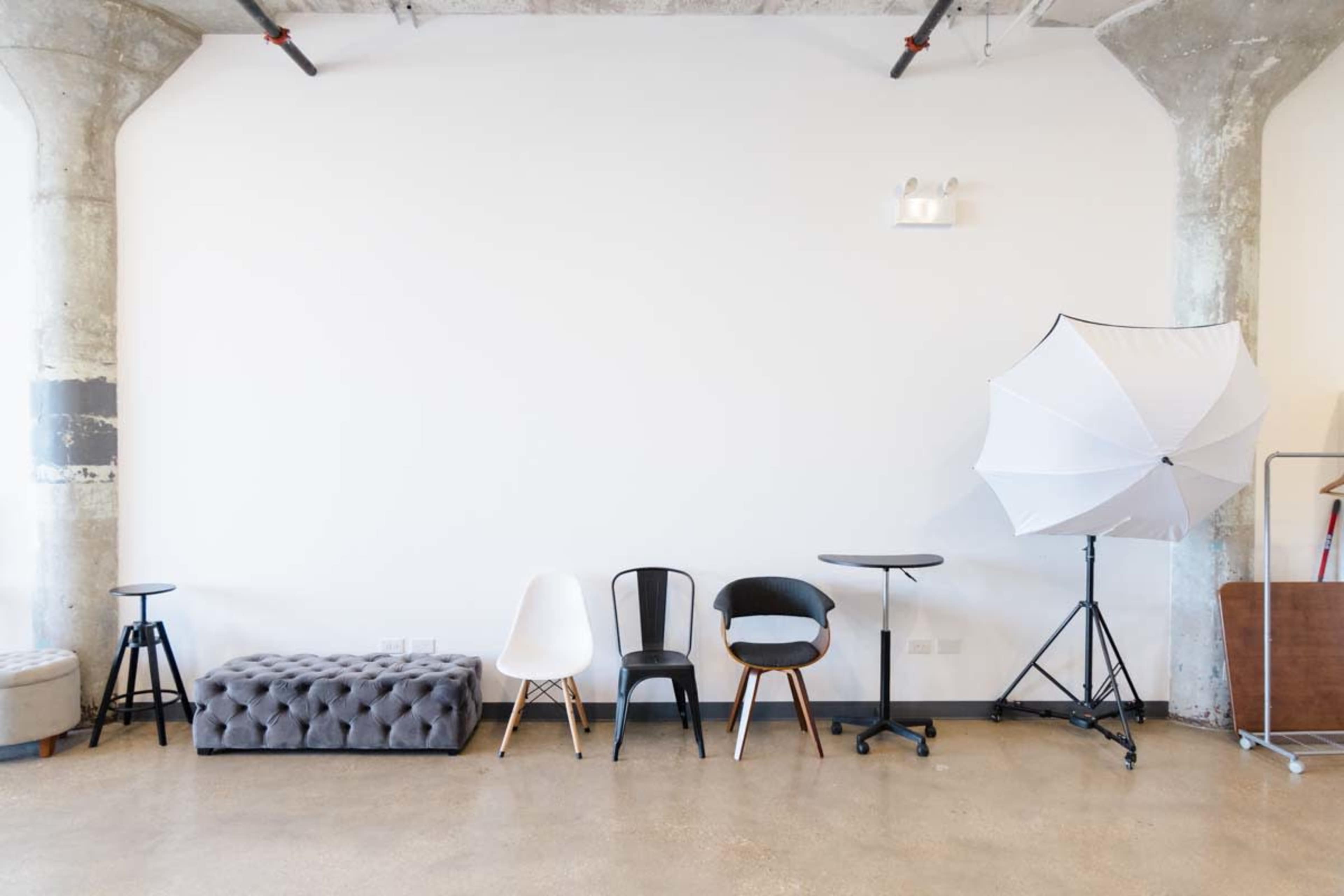 The image shows a minimalist interior featuring various chairs and a photography umbrella stand against a plain wall.
