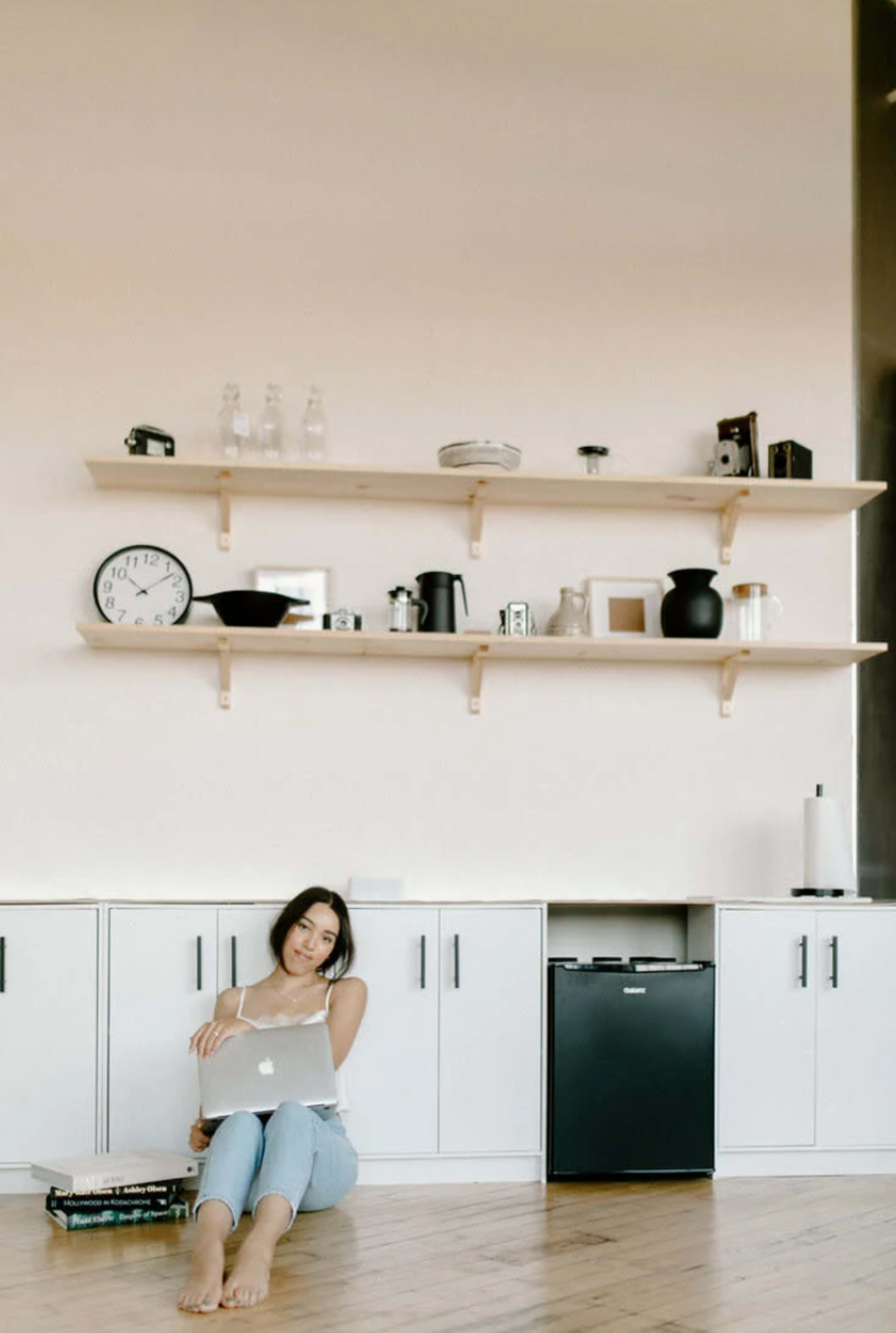 A woman sits on the floor with a laptop in a minimalist room featuring wooden shelves and various household items.