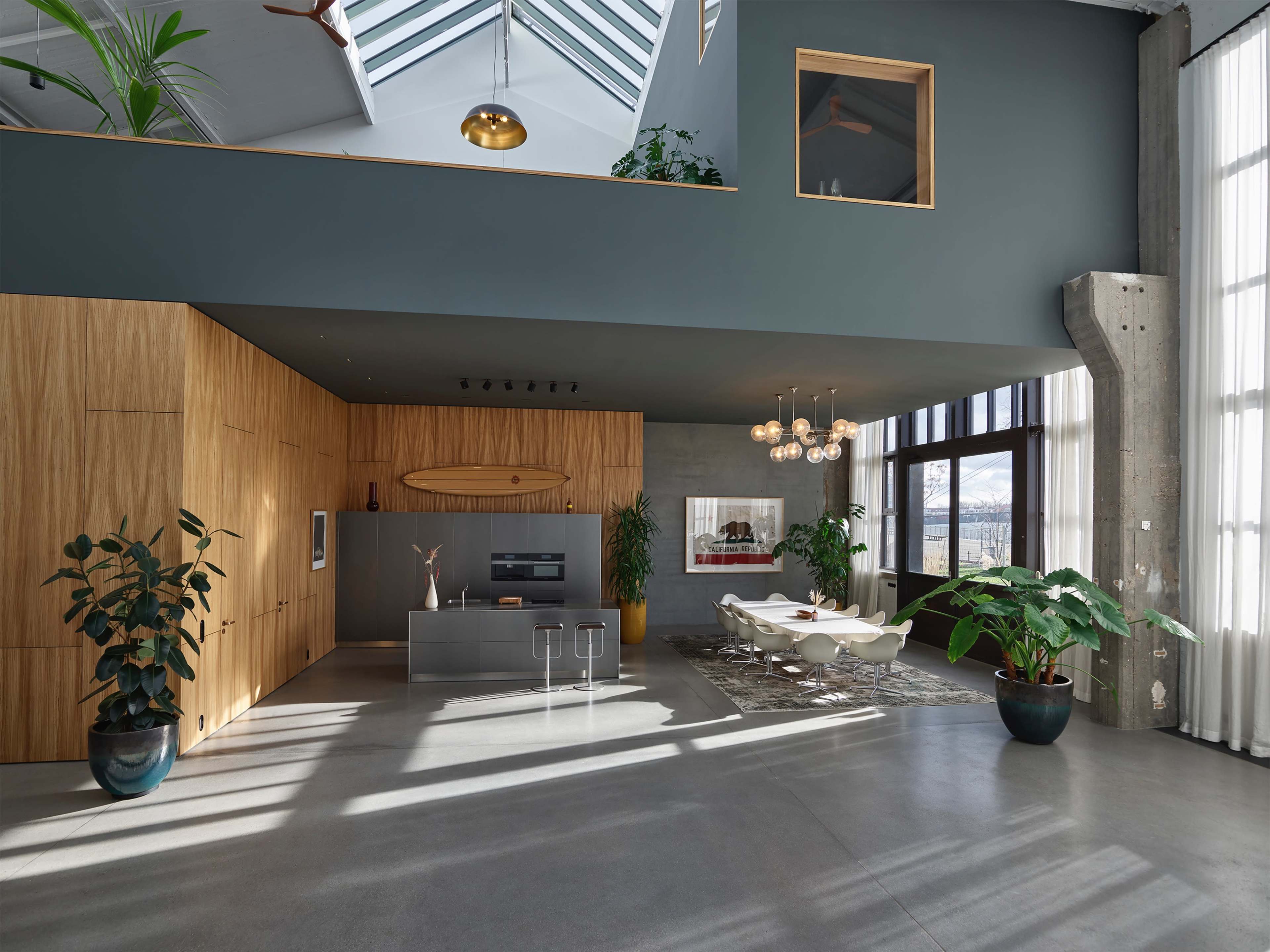 A modern, spacious living area featuring wooden cabinetry, large windows, and contemporary furnishings, with plants positioned throughout the space.
