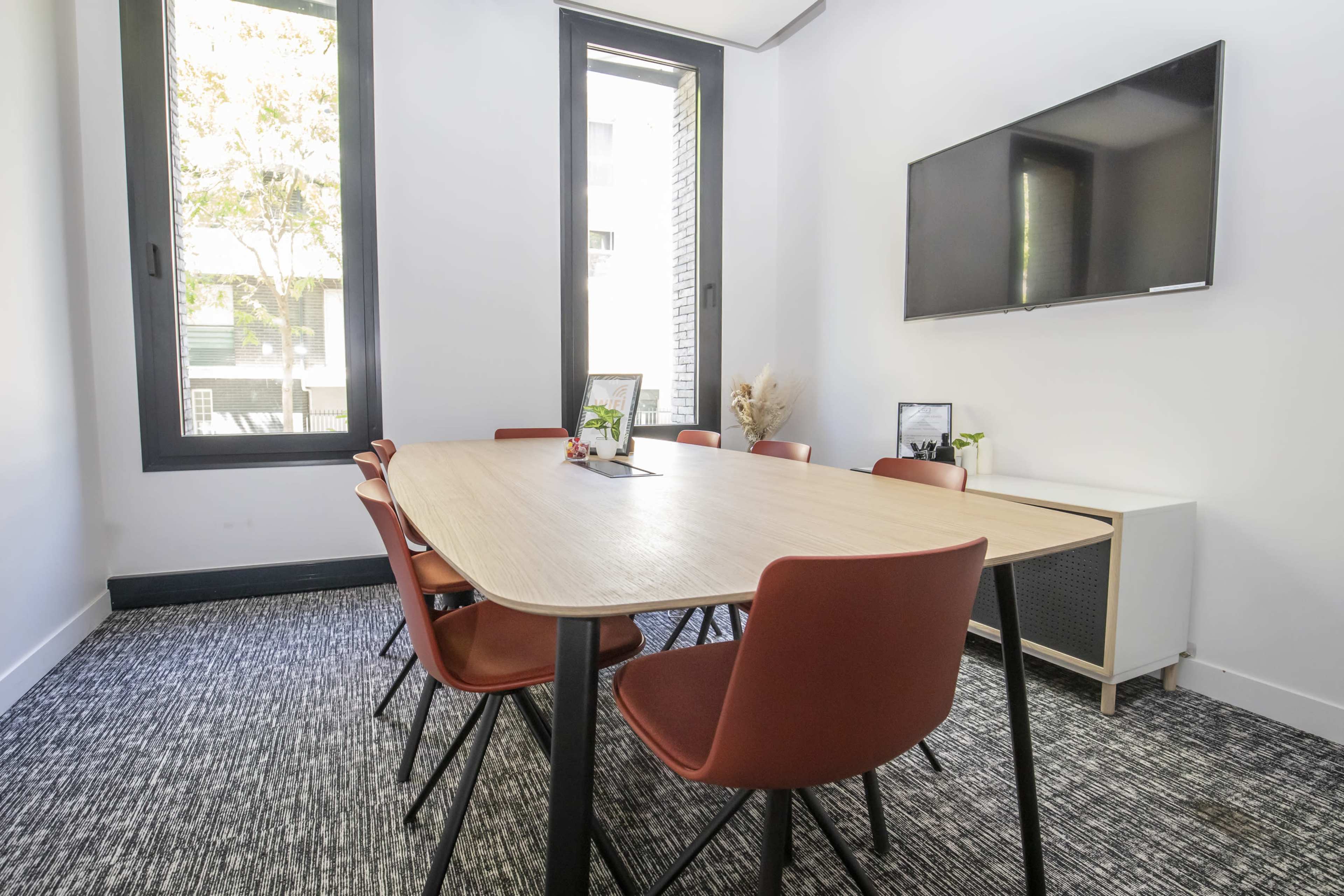 A modern meeting room features a large wooden table surrounded by eight chairs, with large windows allowing natural light to illuminate the space.