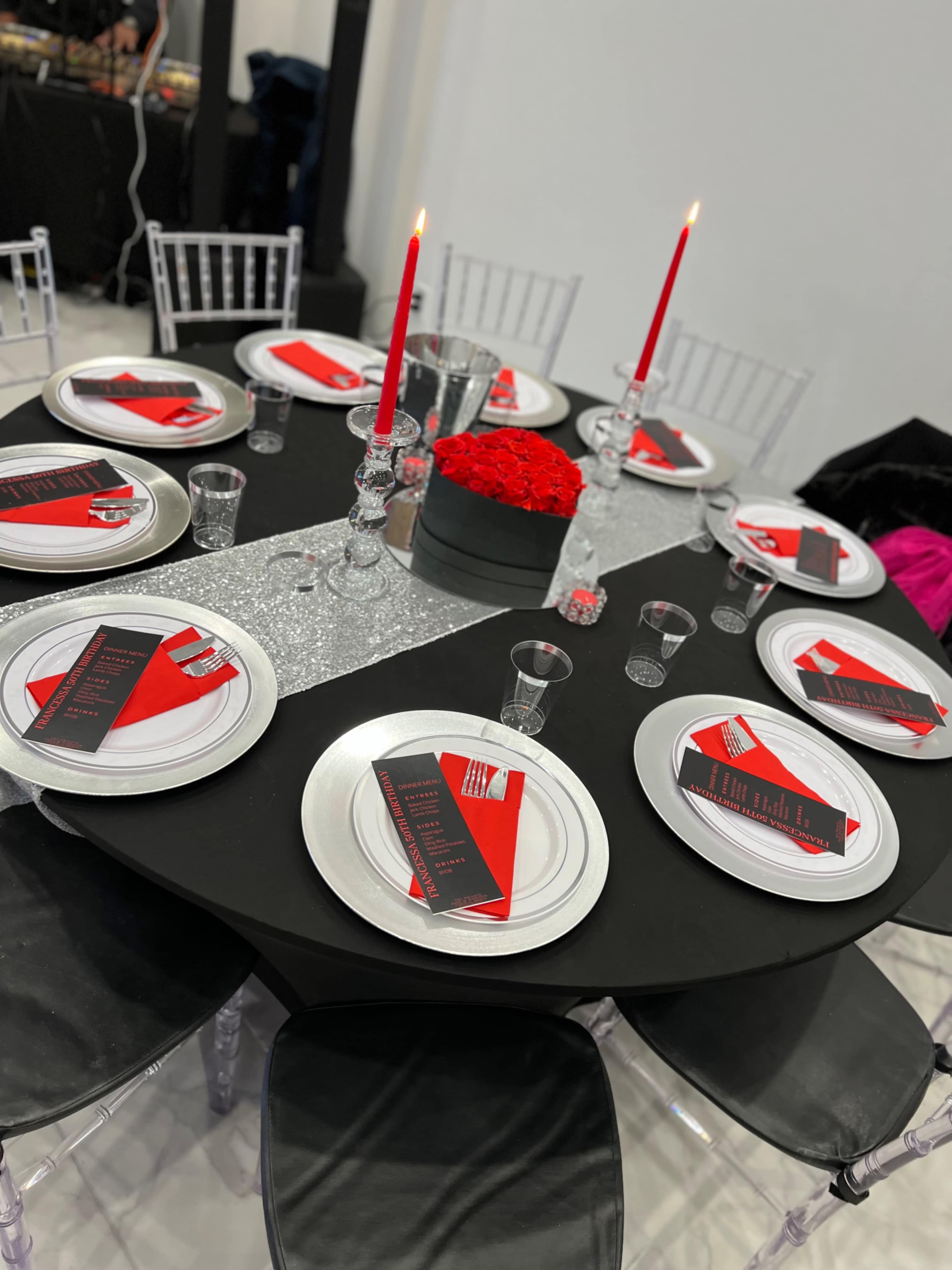 A round dining table is set with black and red themed place settings, silver cutlery, glassware, and two red candles in a floral centerpiece.