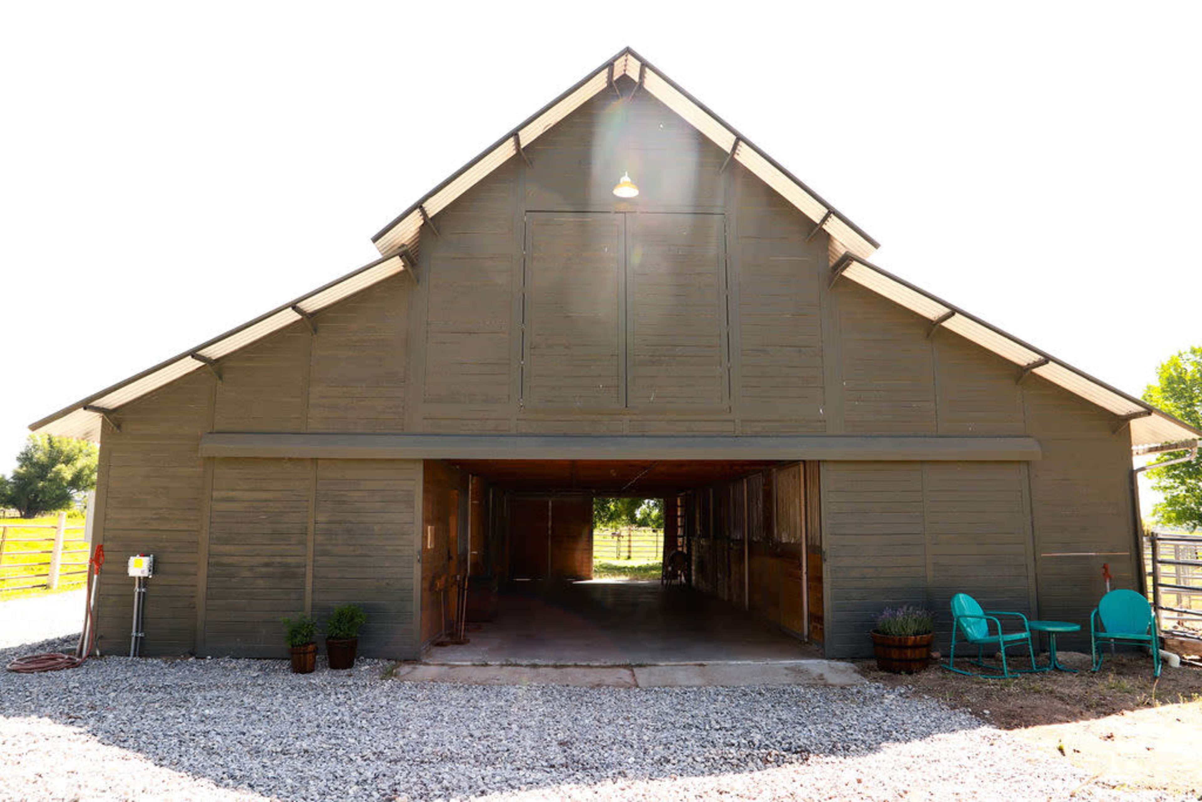 A large, wooden barn with an open front and a gravel yard features two blue chairs on either side.