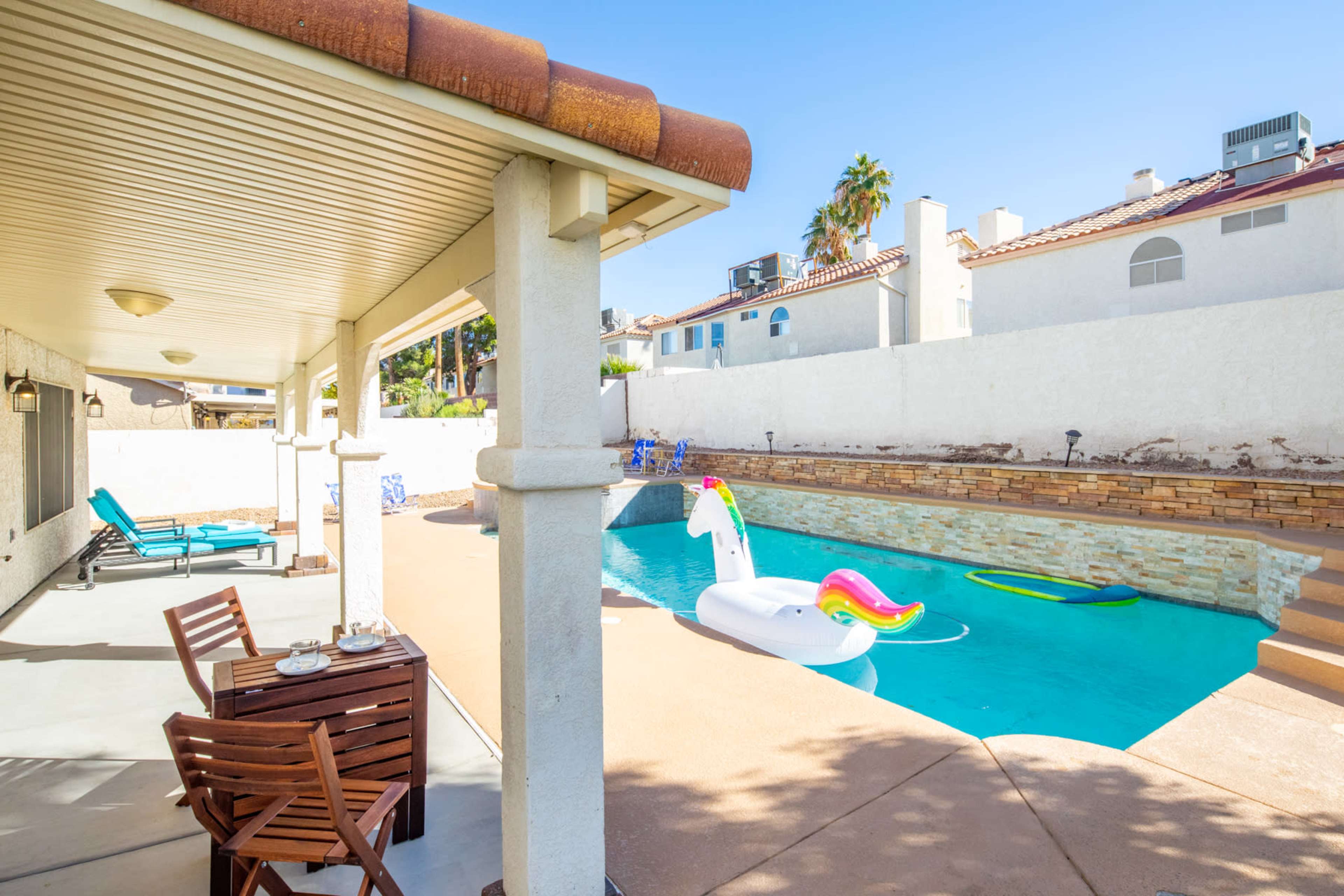 A residential backyard with a swimming pool featuring a unicorn float, surrounded by lounge chairs and a shaded patio area.