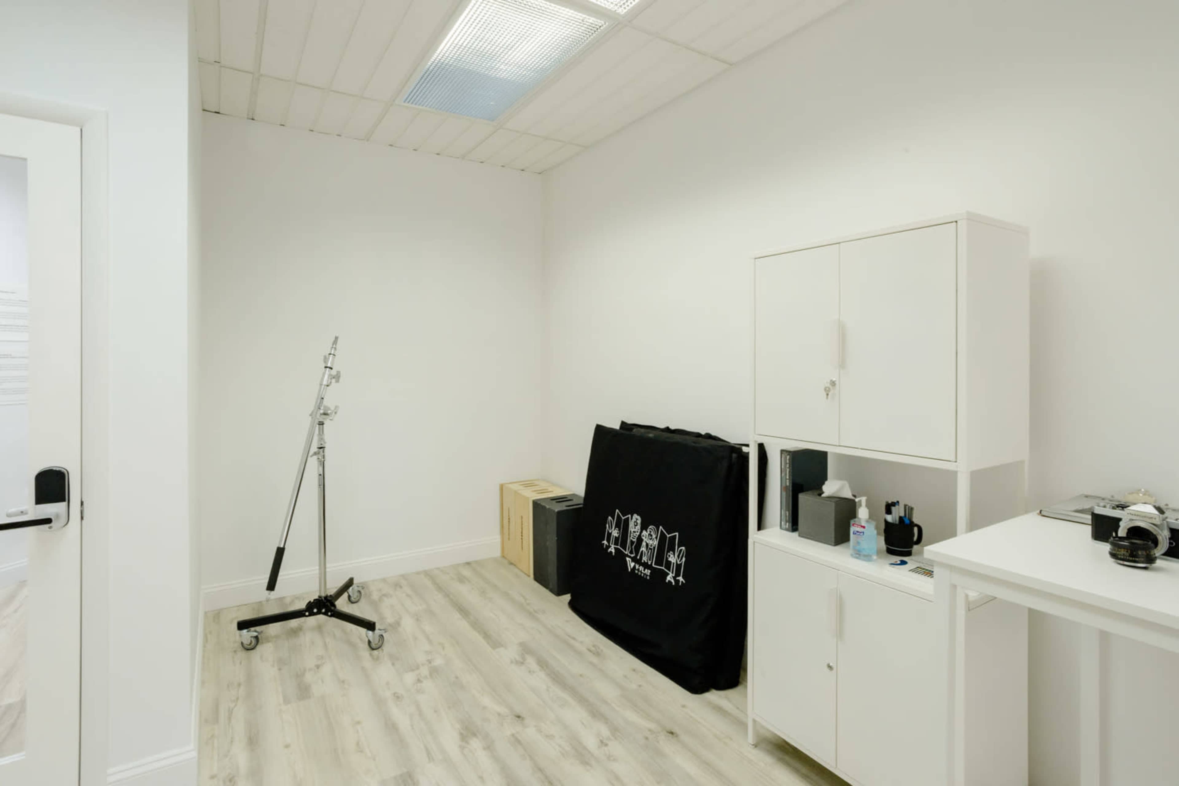 The image shows a minimalistic room with light wood flooring, a closet-like cabinet, a black fabric covering a stand, and a white table with various items on it.