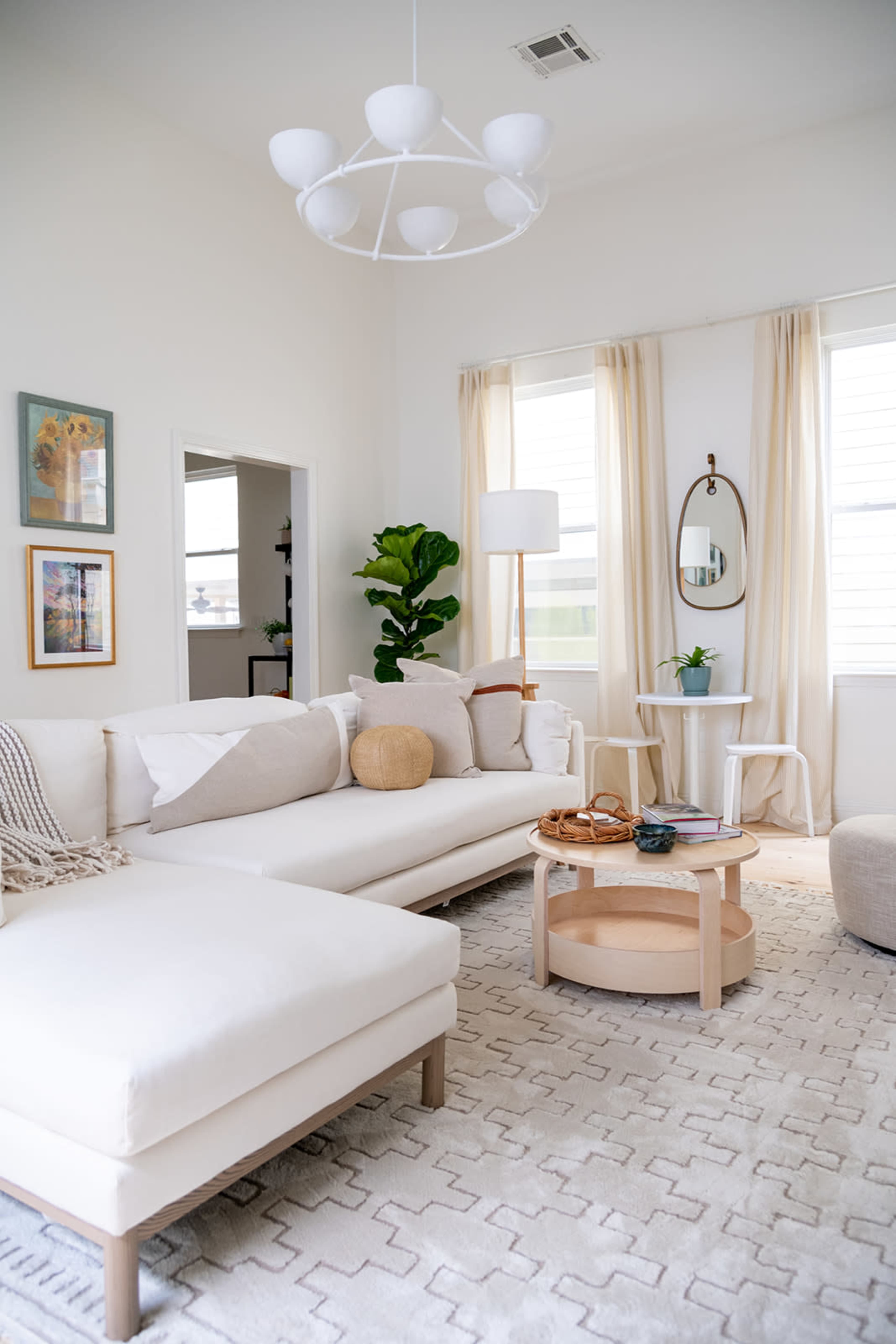 A bright living room features a white sectional sofa, a round coffee table, and a floor lamp, with large windows allowing natural light to illuminate the space.