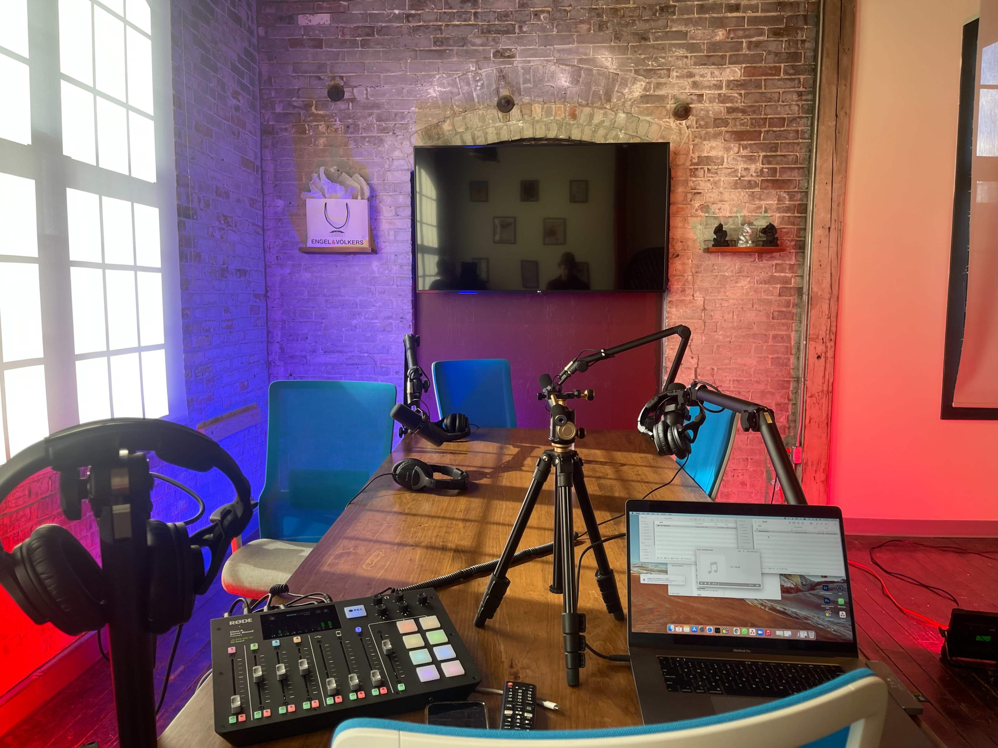 A podcast recording studio features a wooden table with microphones, headphones, a laptop, and a television on a brick wall backdrop illuminated in red and blue lights.