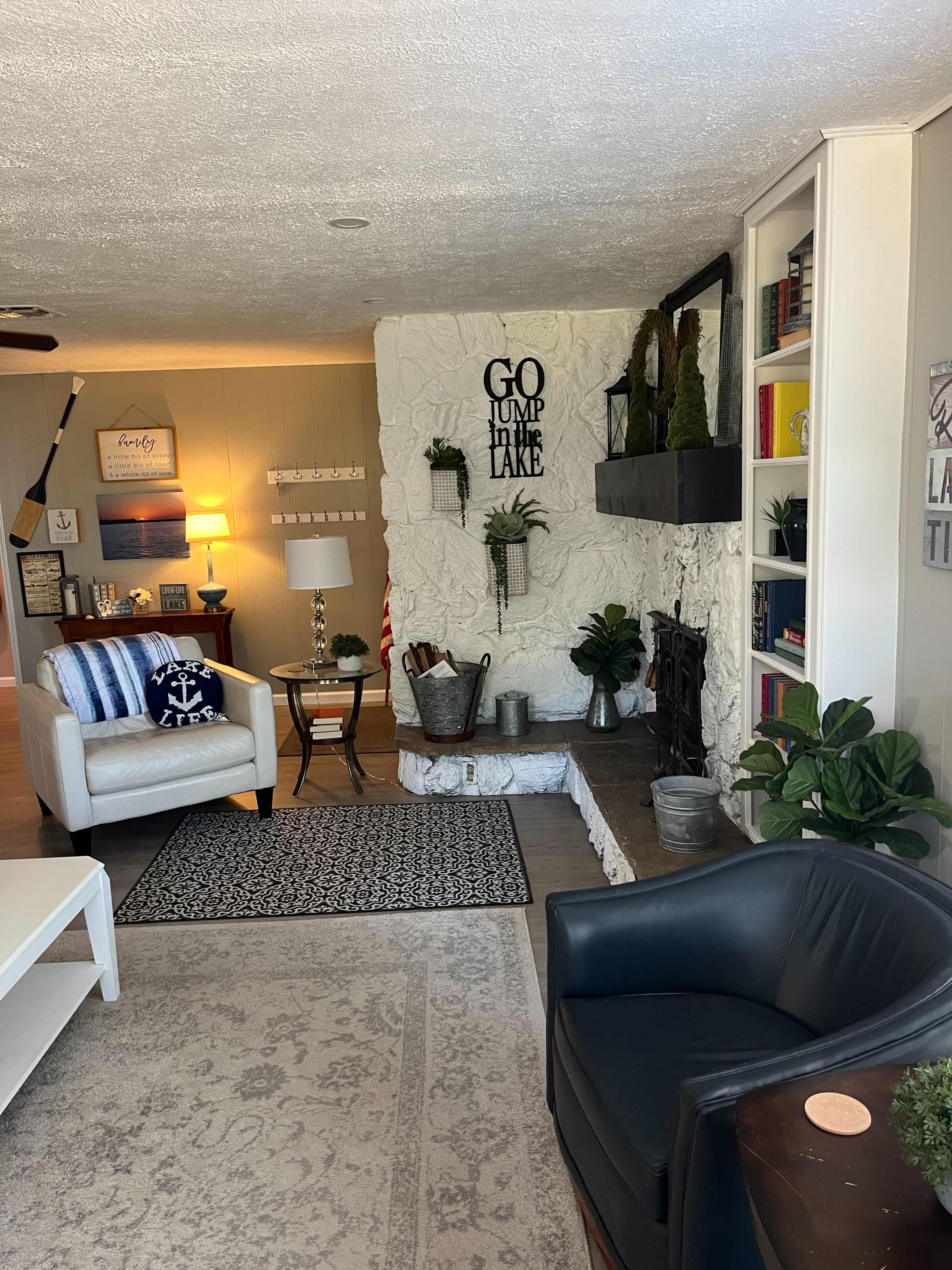 The living room features a cozy seating area with a white chair and a black chair, a stone fireplace, and decorative plants on shelves and tables.