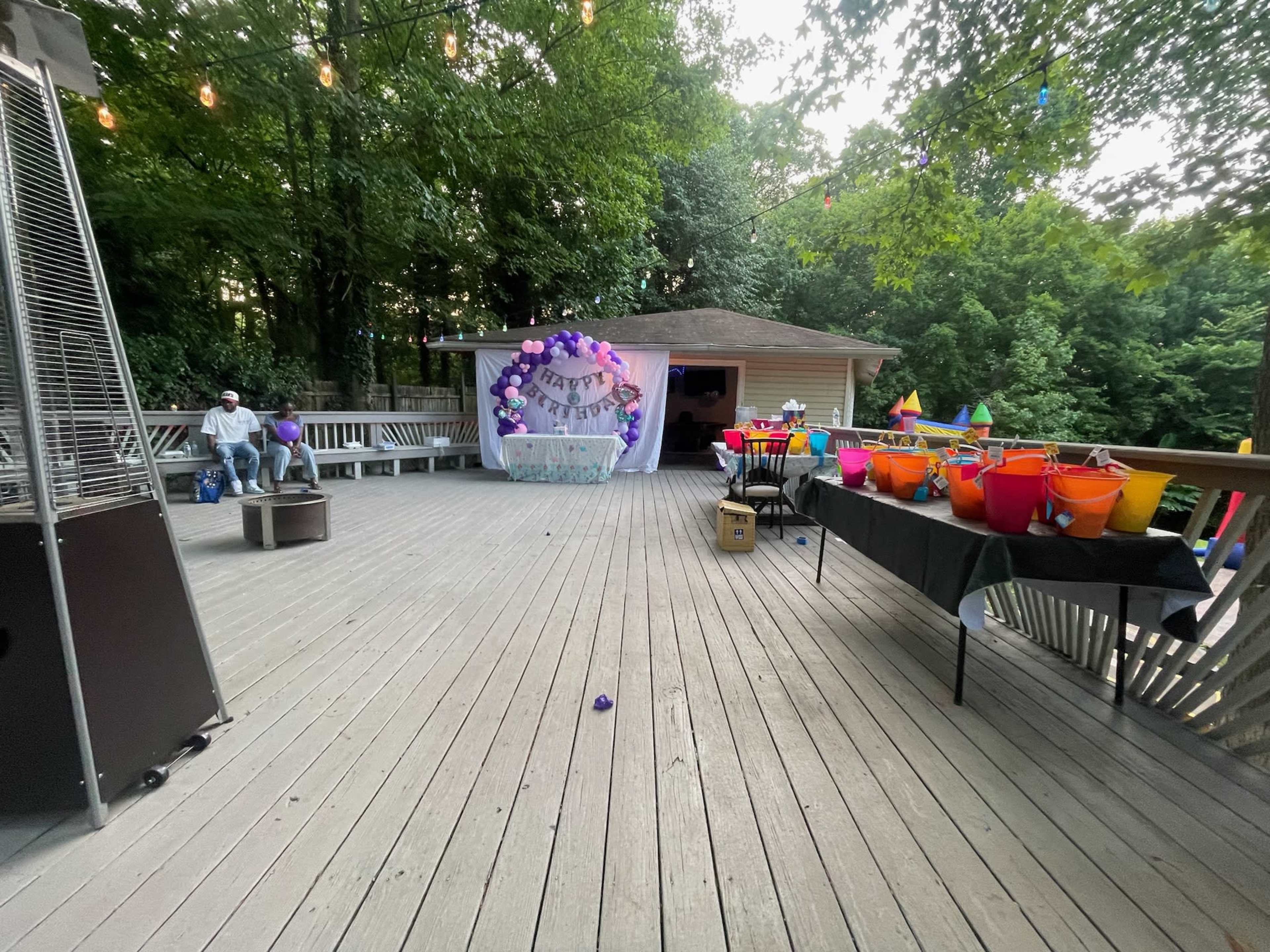 A decorated outdoor deck features a backdrop with "Happy Birthday," colorful party buckets lined on a table, and string lights overhead.
