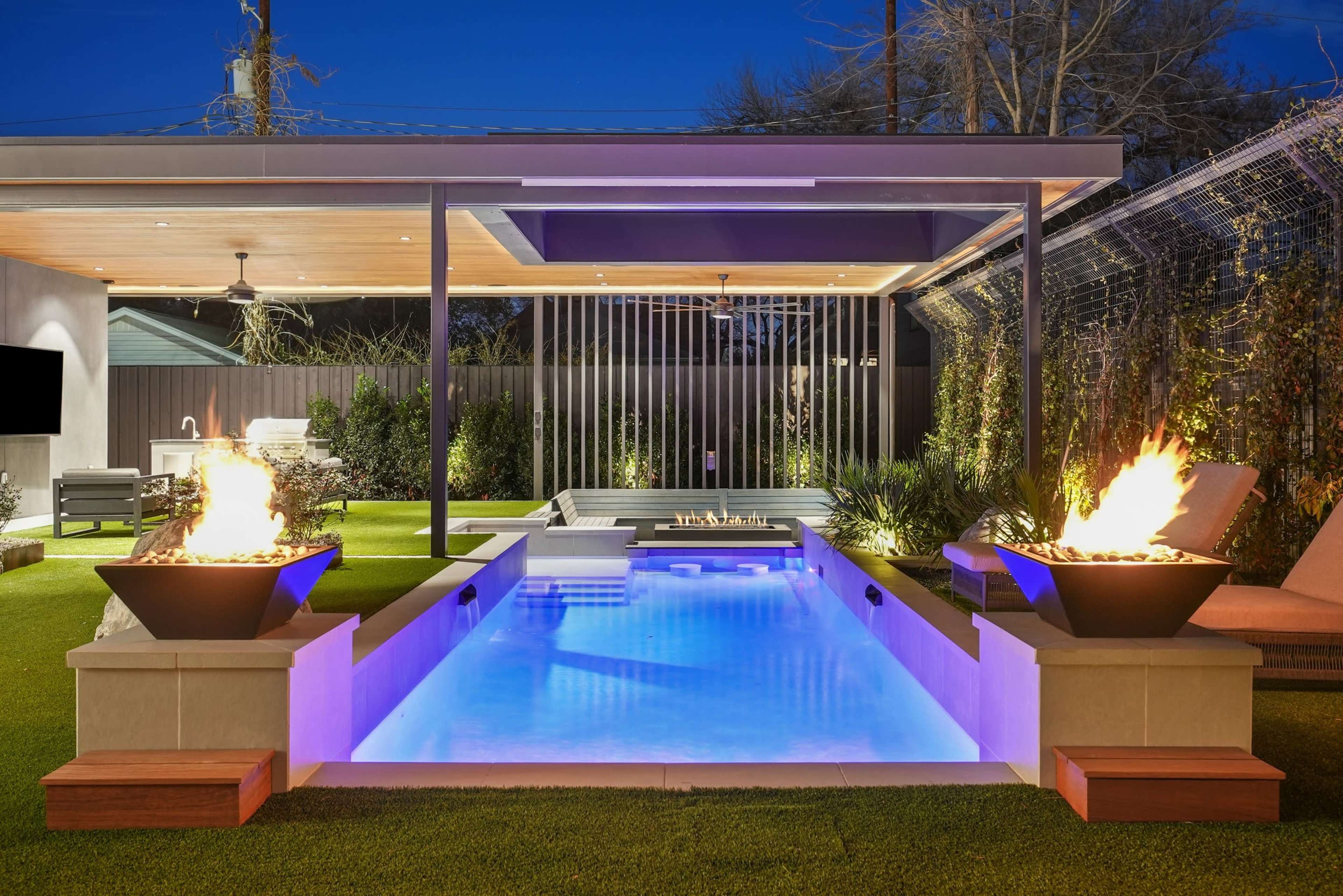 The image depicts a modern backyard with a swimming pool illuminated by flames in two planters, framed by a covered seating area and landscaped greenery under a twilight sky.