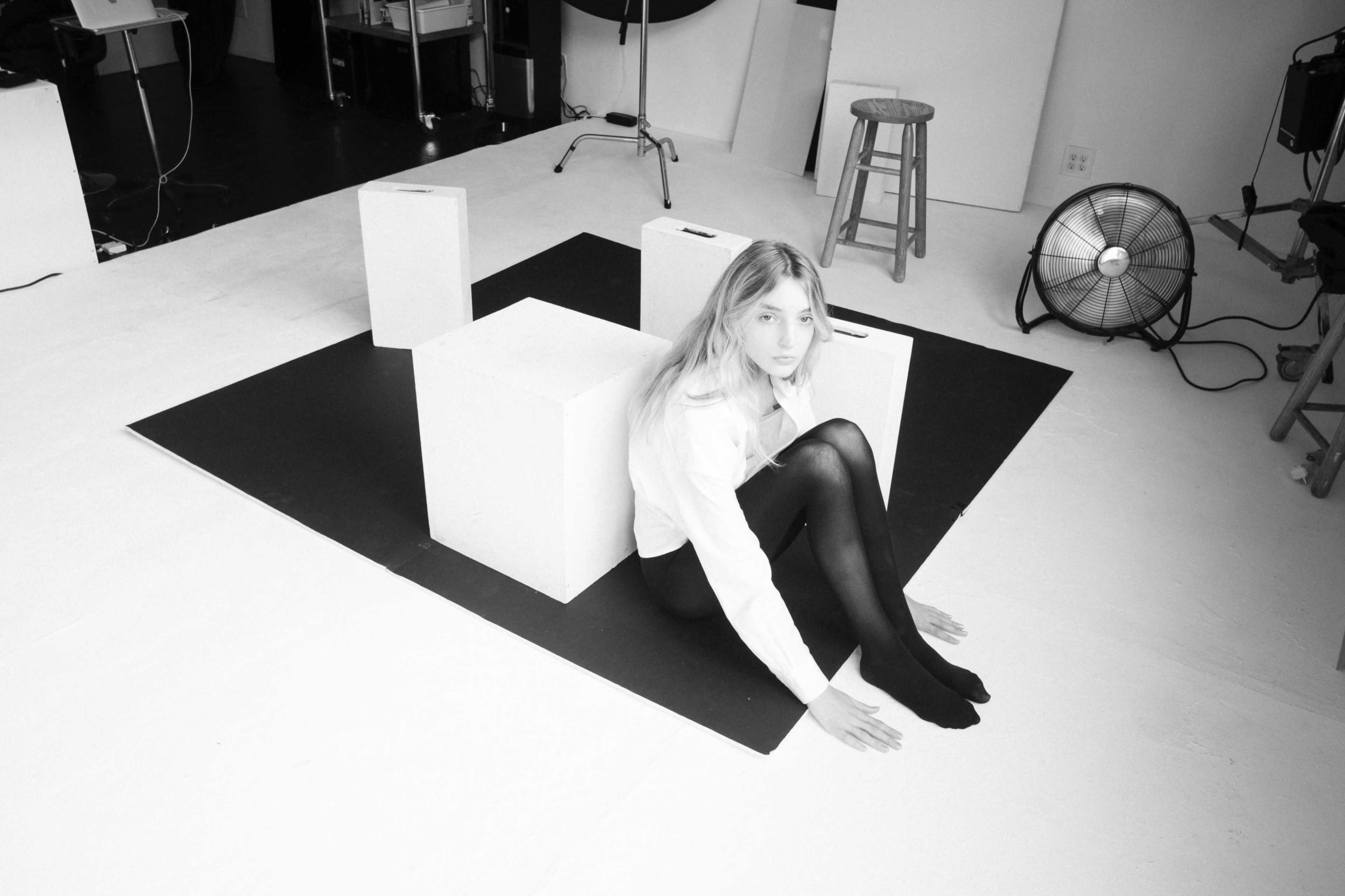 A woman sits on a black mat surrounded by white pedestals in a studio space.