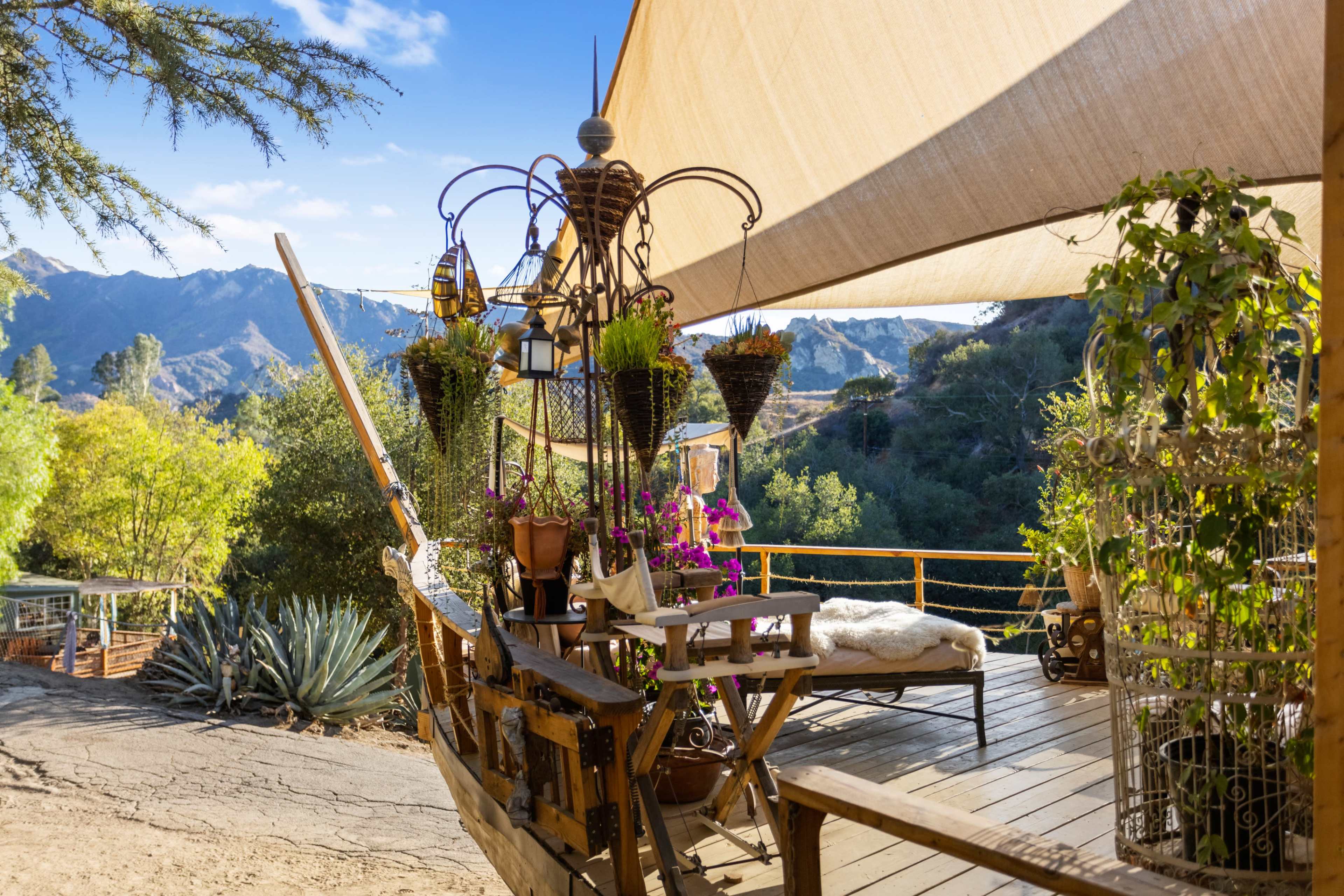 A wooden deck features a boat structure adorned with potted plants and lanterns, overlooking a mountainous landscape under a shaded canopy.