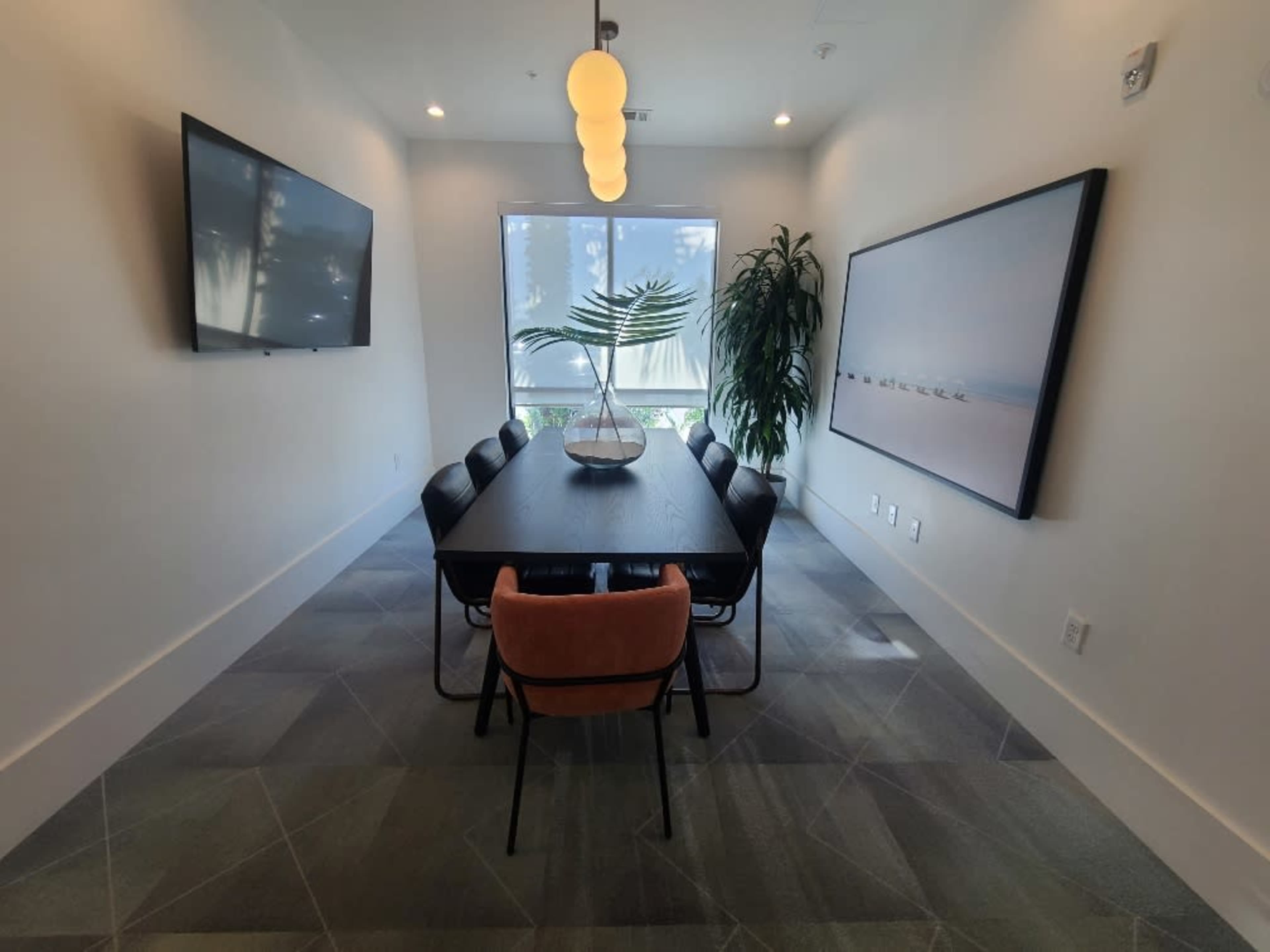 A modern conference room features a long table surrounded by chairs, with a TV on the wall and a large window letting in natural light.