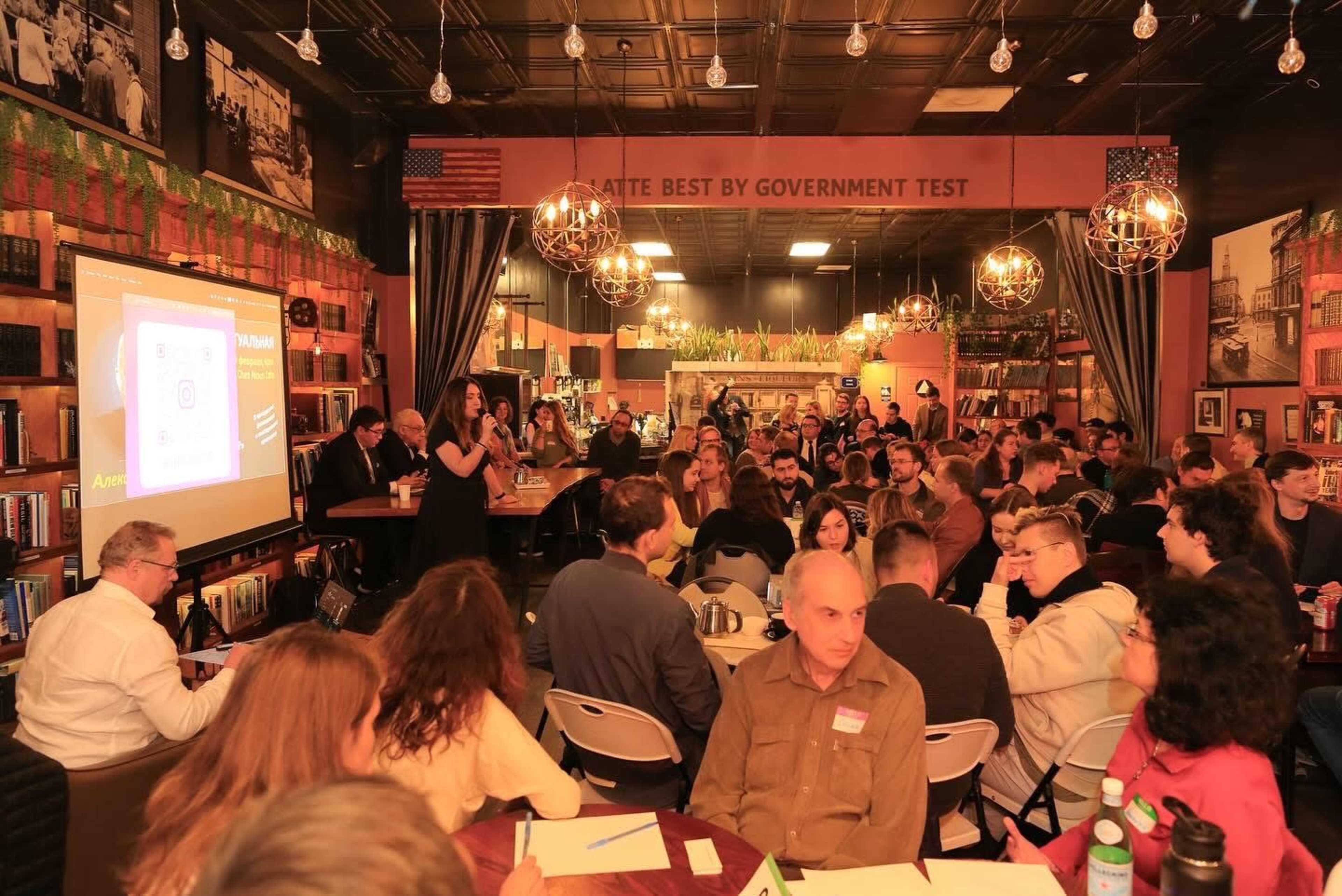 A large group of people is seated in a café setting, engaging in a presentation while others participate in discussions around tables.