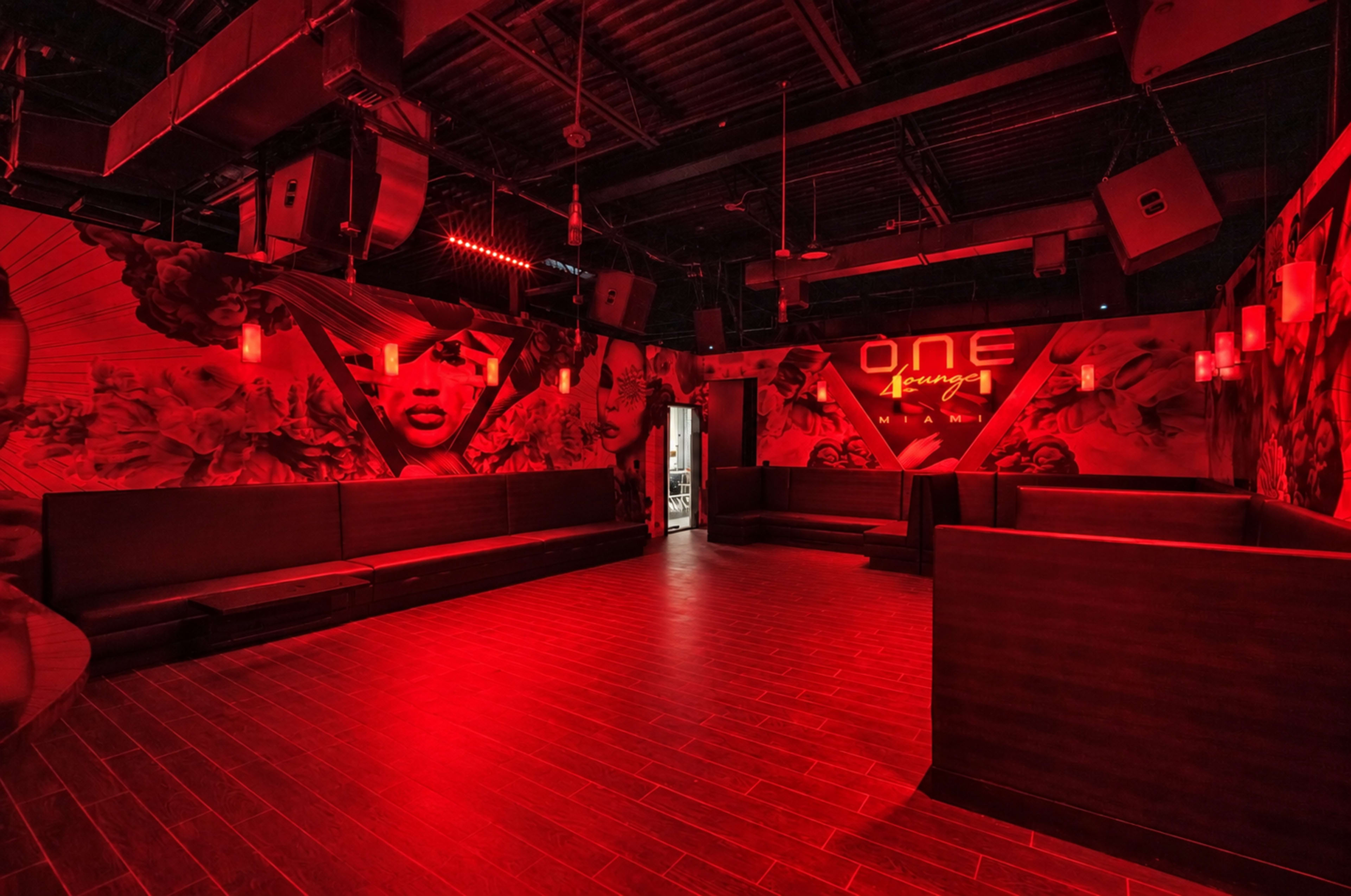 The image shows an empty nightclub interior featuring red lighting and a mural on the wall, with several seating areas arranged along the perimeter.