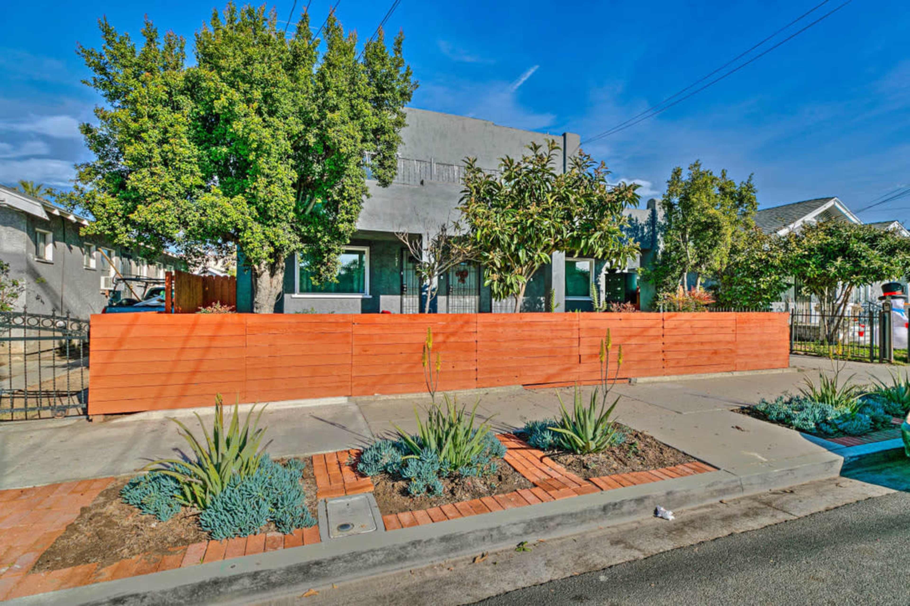 The image shows a modern gray house with a distinctive wooden fence and landscaped garden, situated on a quiet street.
