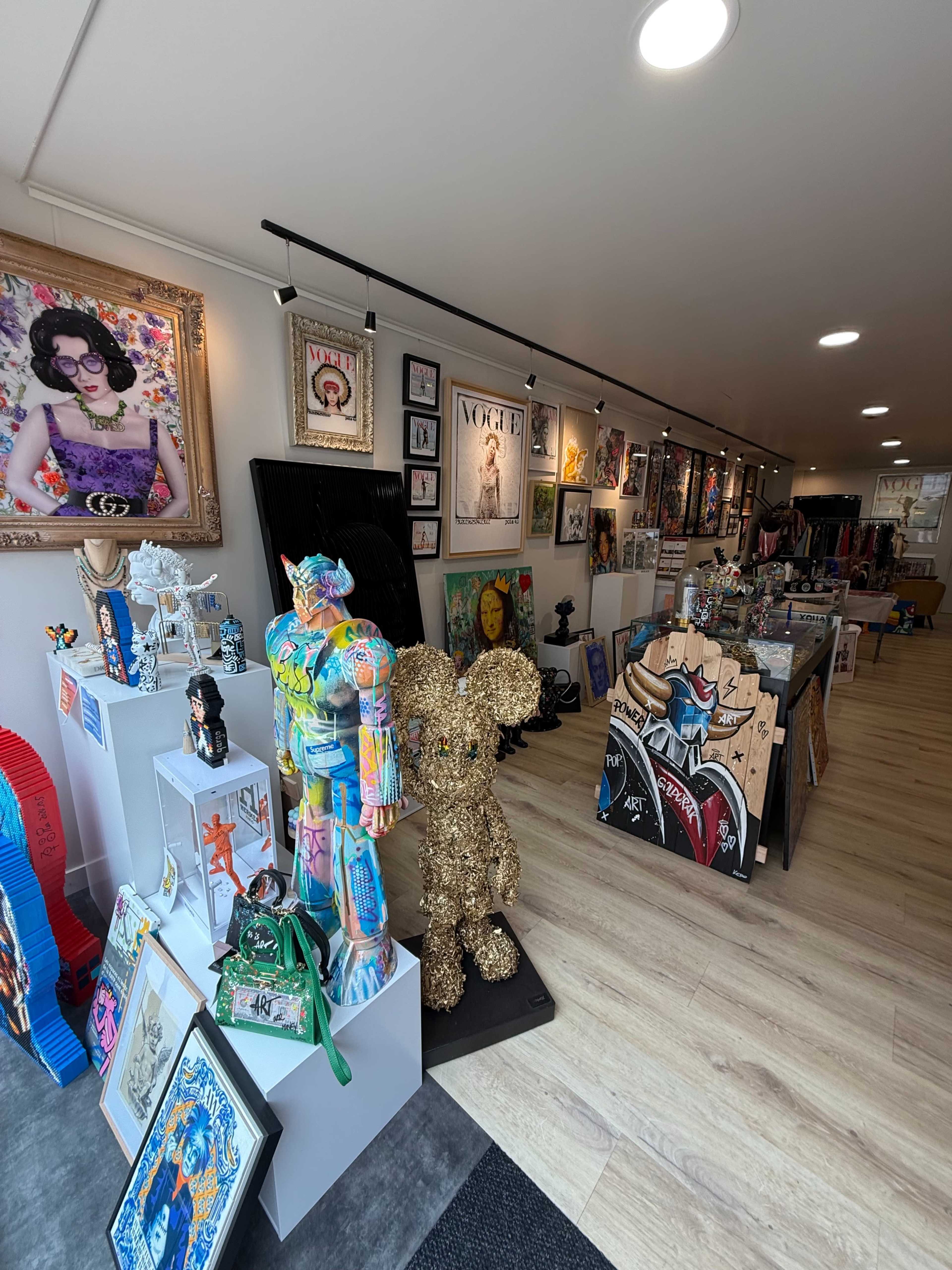 The image shows a colorful art gallery featuring various artwork and sculptures, with displays along the walls and several pieces arranged on stands and tables.