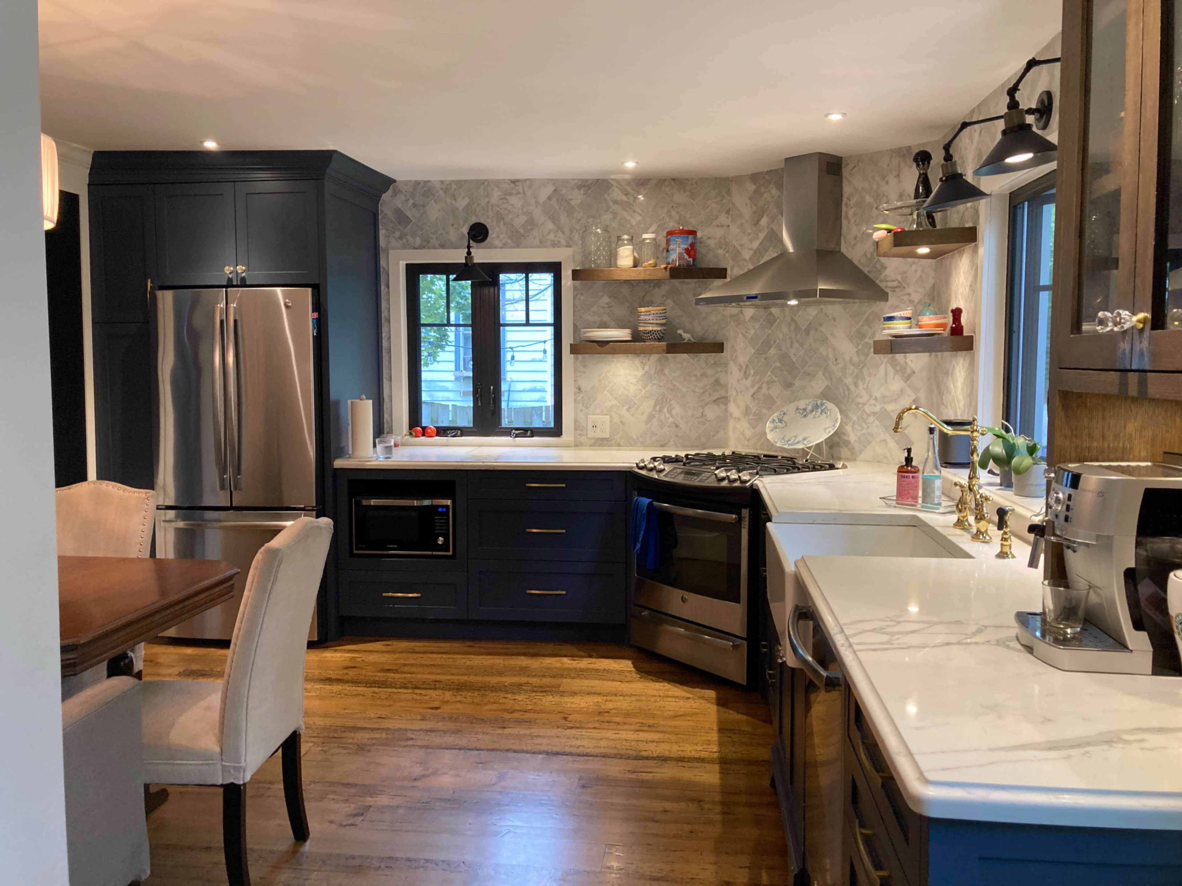 A modern kitchen featuring dark blue cabinetry, stainless steel appliances, a marble backsplash, and a dining area with a wooden table and upholstered chairs.