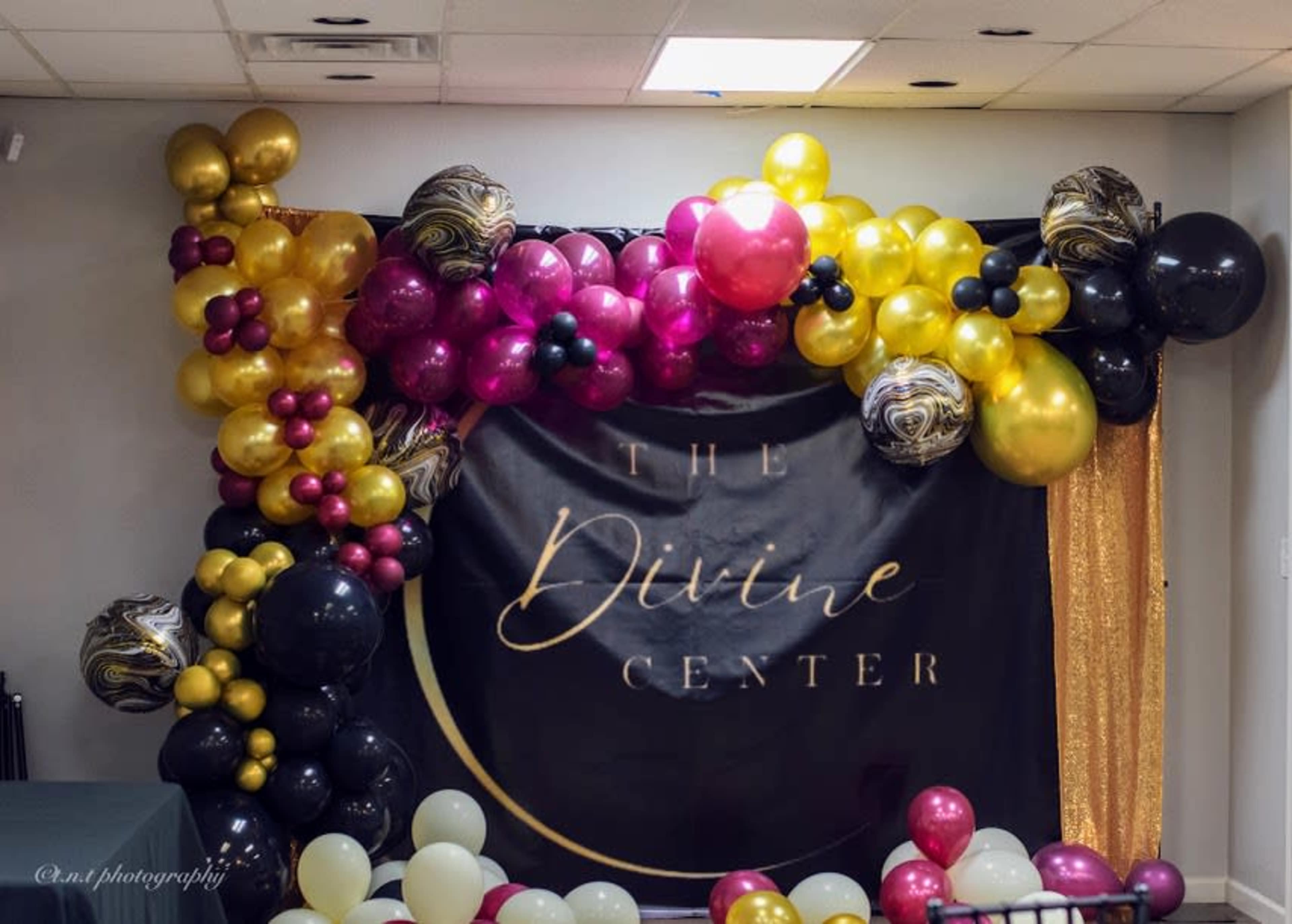 A decorative backdrop made of balloons in various colors, including gold, black, and burgundy, with the text "The Divine Center" prominently displayed in the center.