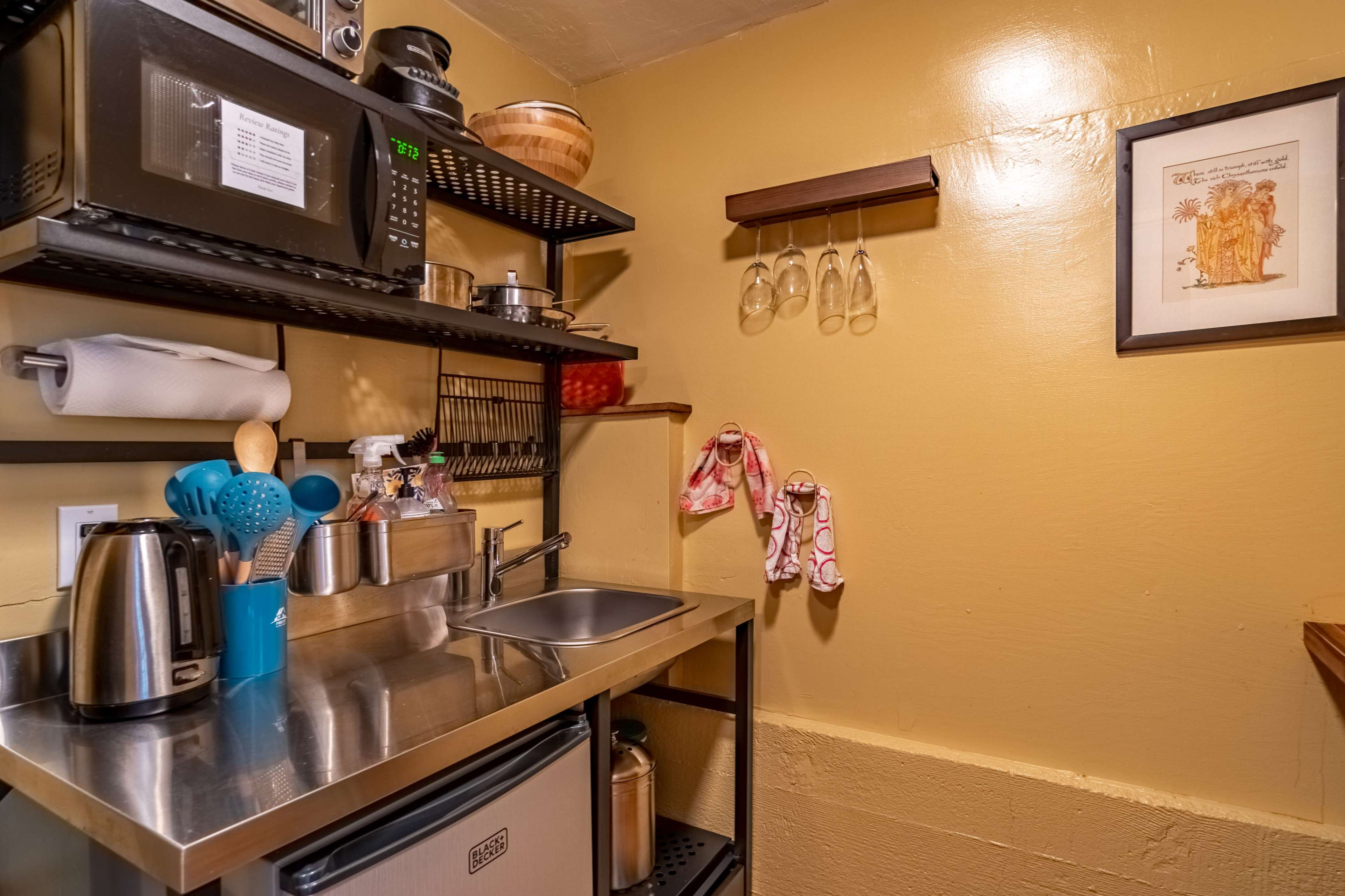 A compact kitchen area featuring stainless steel countertops, a small sink, a microwave, and shelves displaying kitchen utensils and glassware.