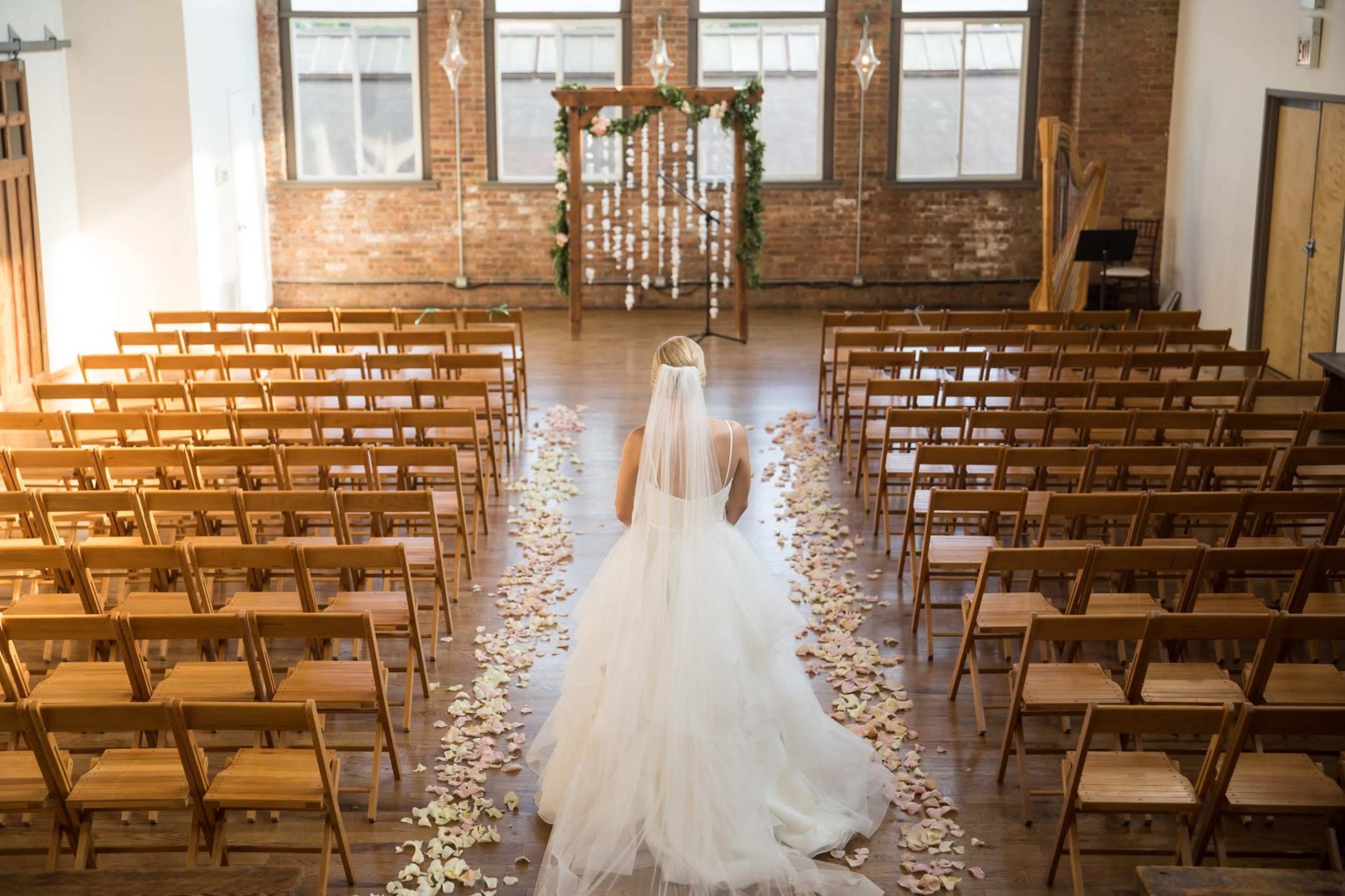 A bride in a white gown stands at the end of a aisle lined with flower petals in a warmly lit ceremony space with wooden chairs and brick walls.