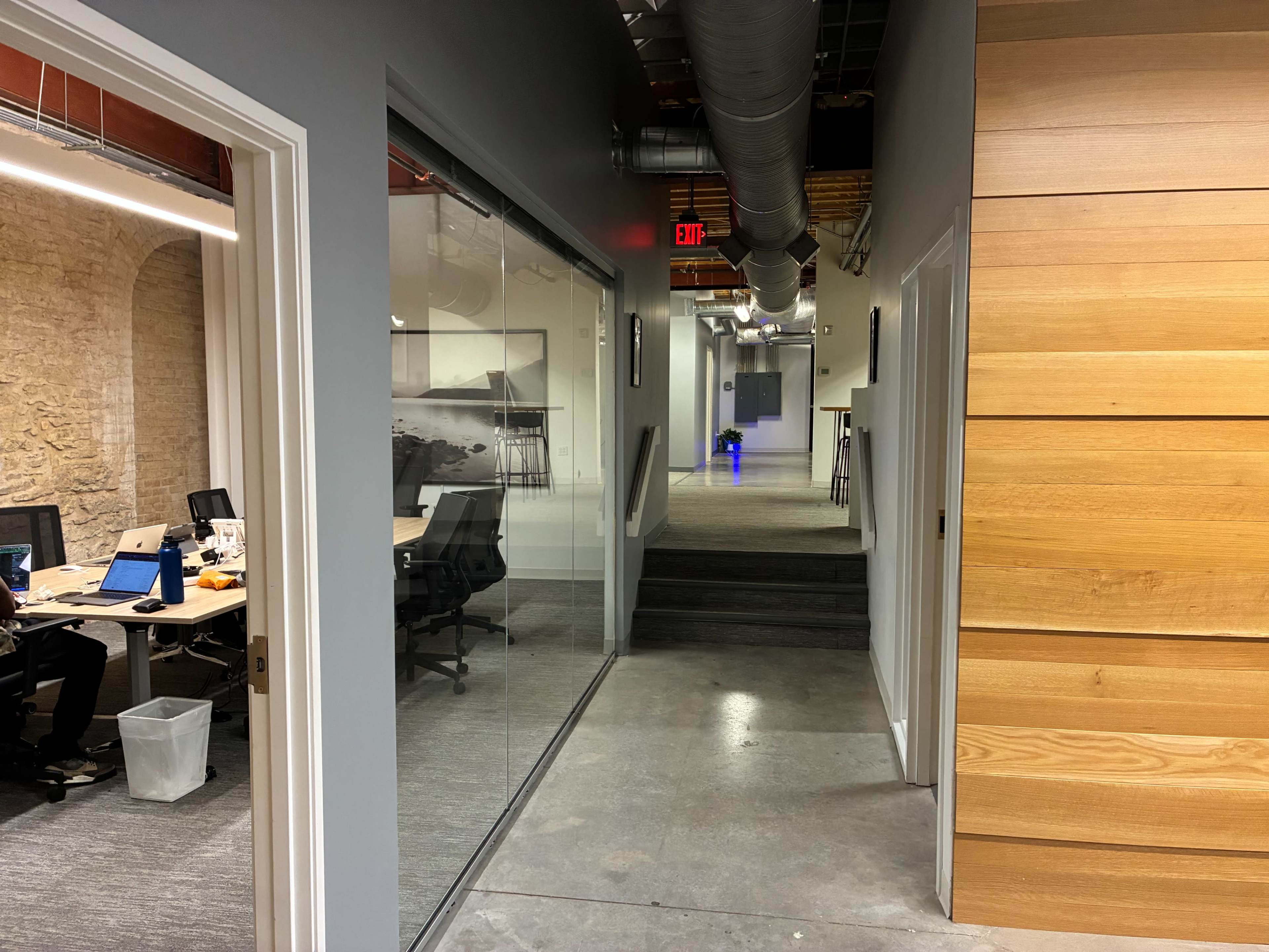 The image shows a modern office corridor with a view of workspace areas on one side and a series of steps leading to a hallway on the other.