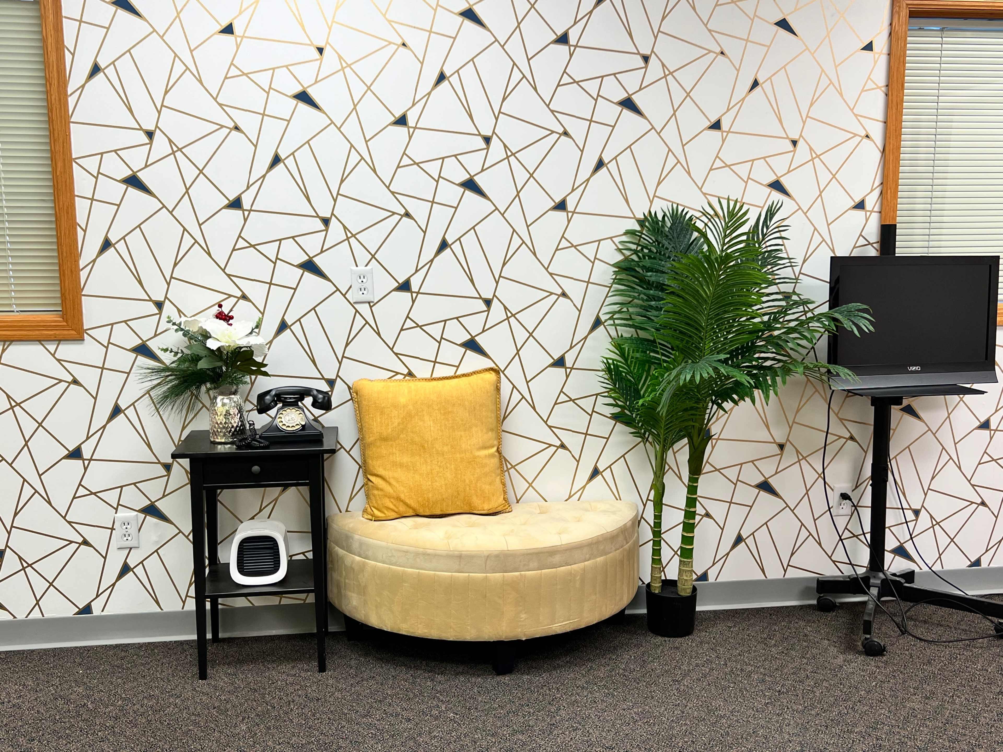A circular yellow seat is placed next to a small black table with a telephone and a decorative plant, set against a geometric patterned wall and accompanied by a tall artificial palm.
