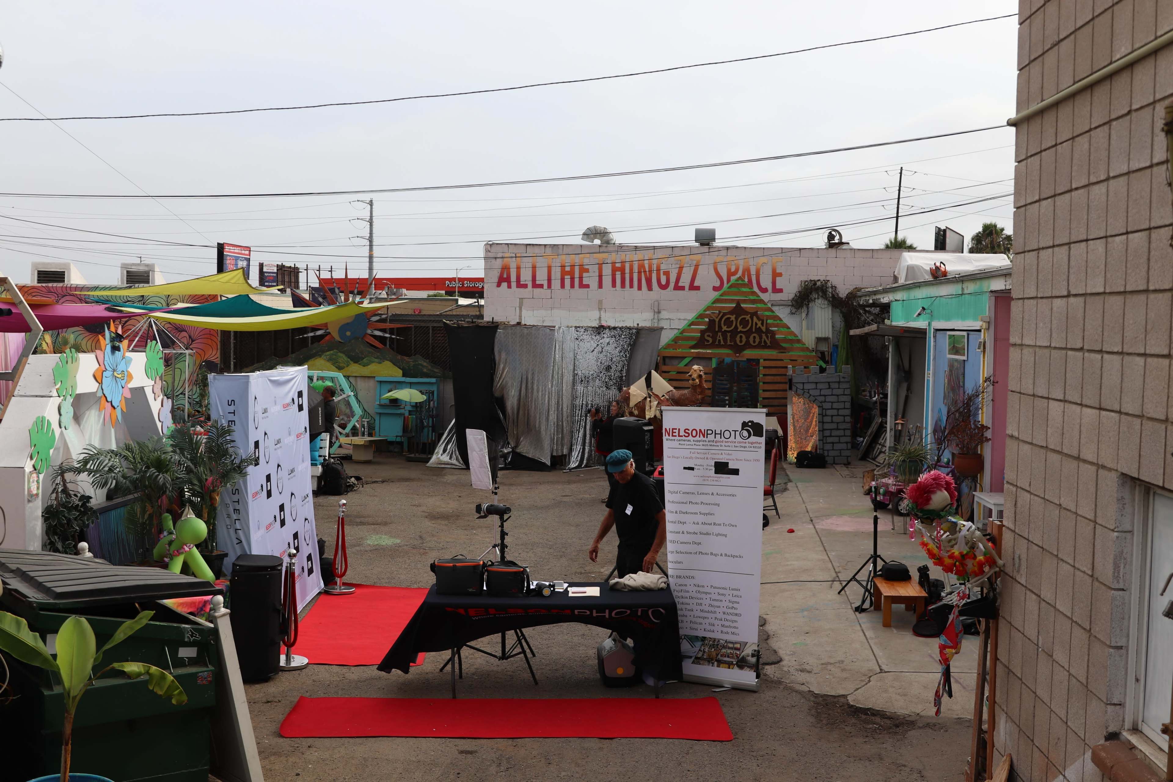 A small outdoor area featuring a black table with a photography setup, flanked by colorful decorations and a mural that says "ALL THINGZ SPACE."