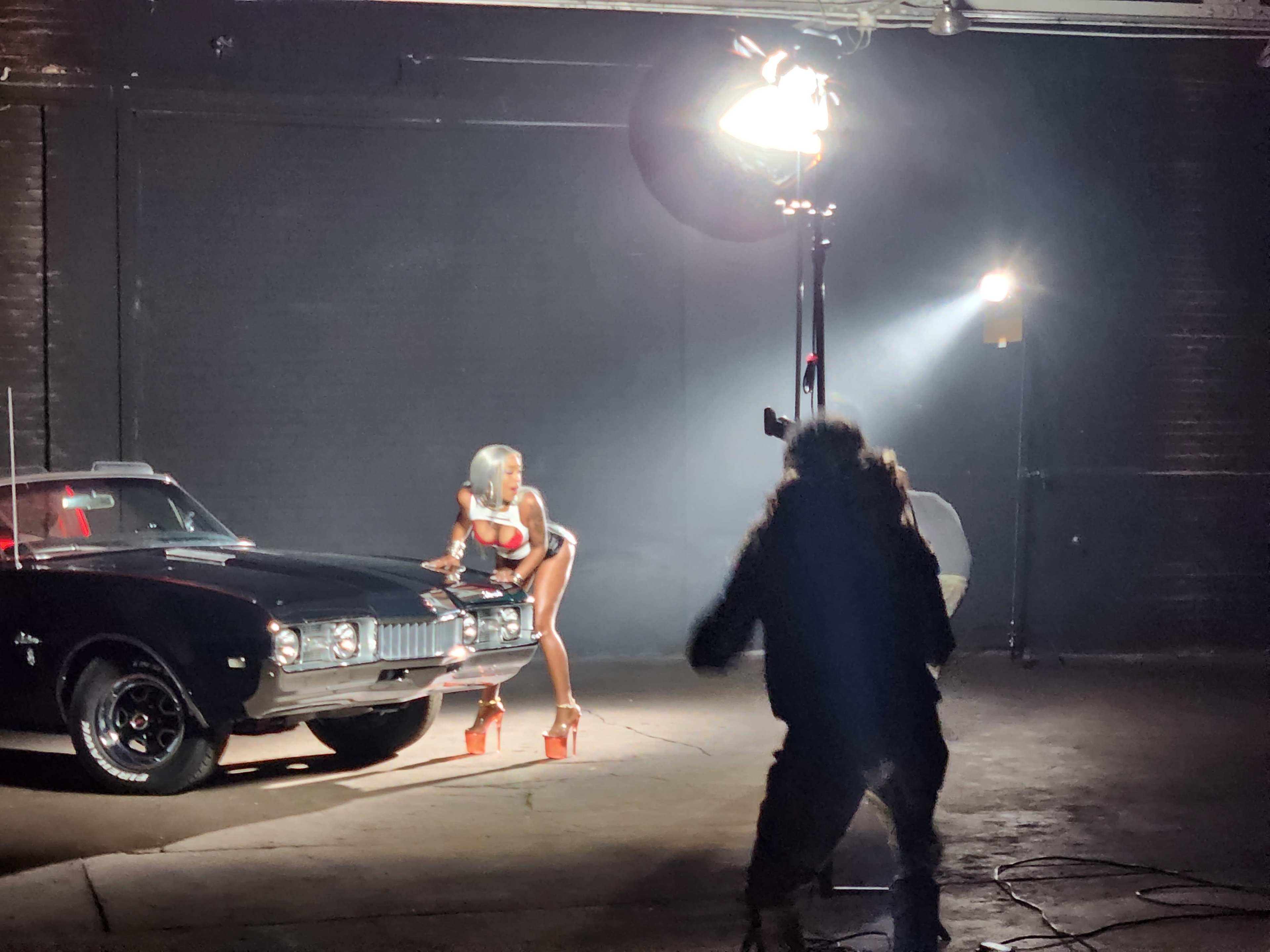 A person poses beside a classic black car while a photographer captures the scene under bright studio lights.