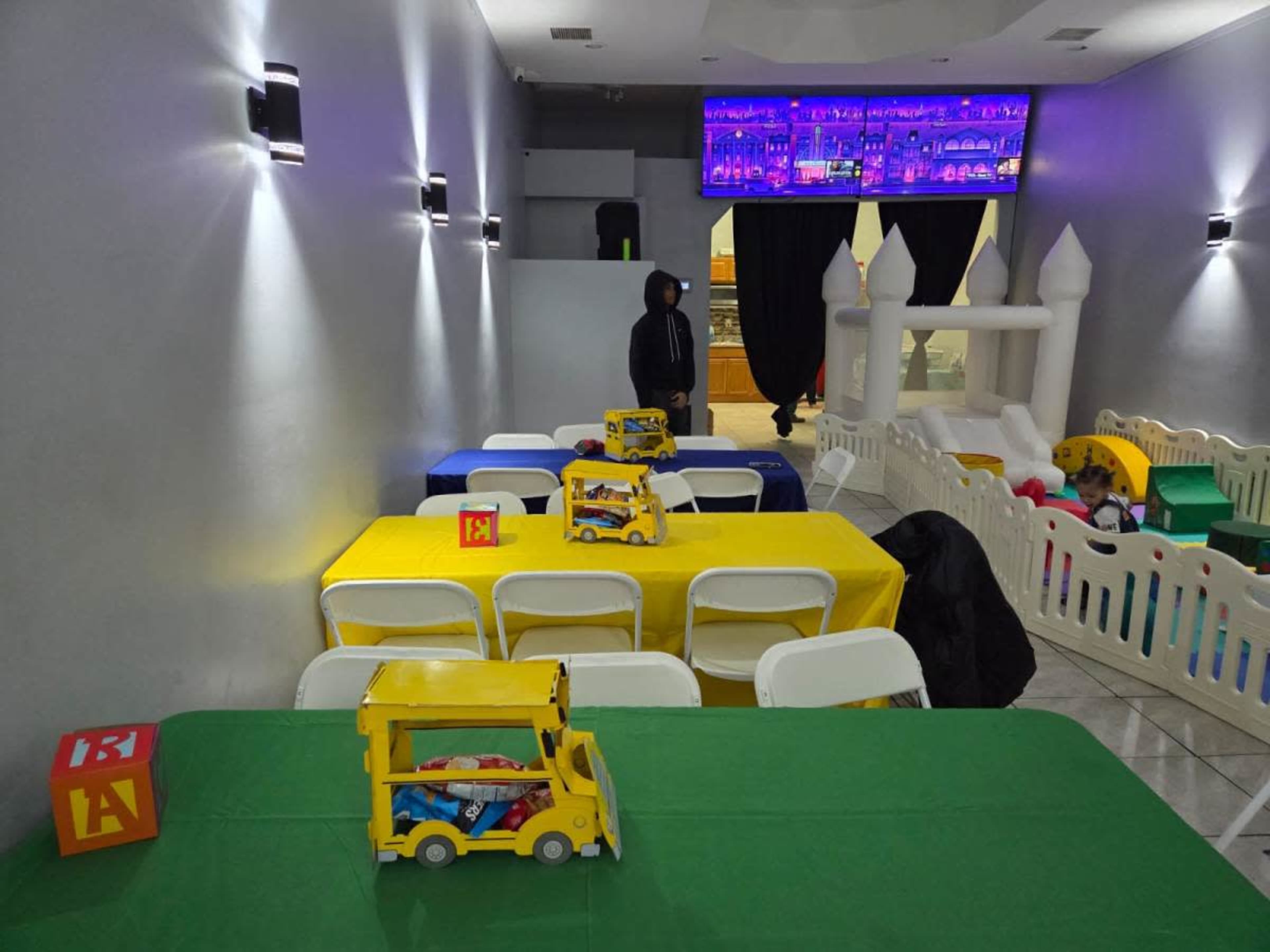 The image shows a brightly colored children's party space with tables covered in blue, yellow, and green cloths, and a play area with toys in the background.