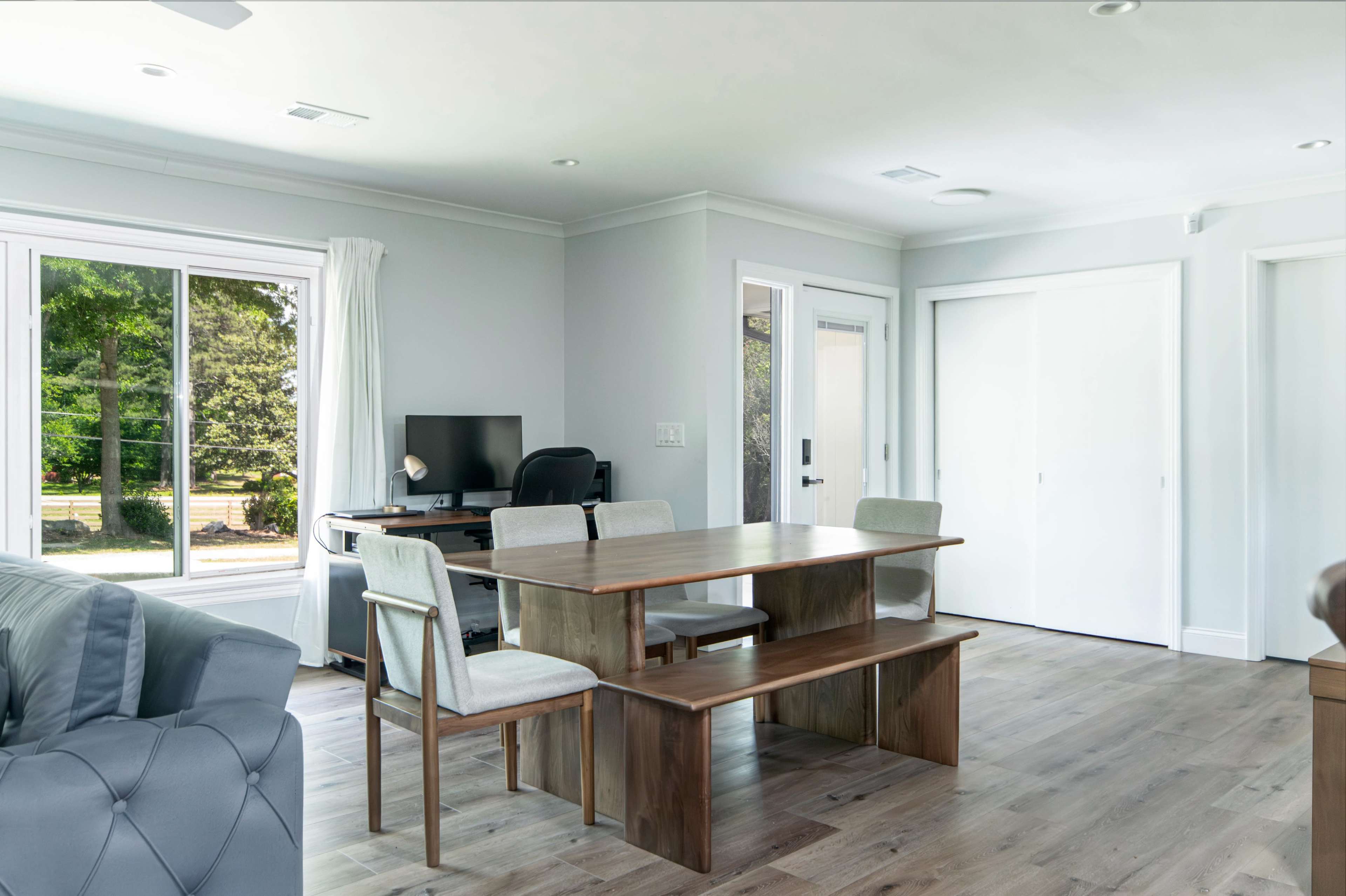 The image shows a modern living space with a wooden dining table and four chairs, a computer desk with a chair, and large windows allowing natural light.