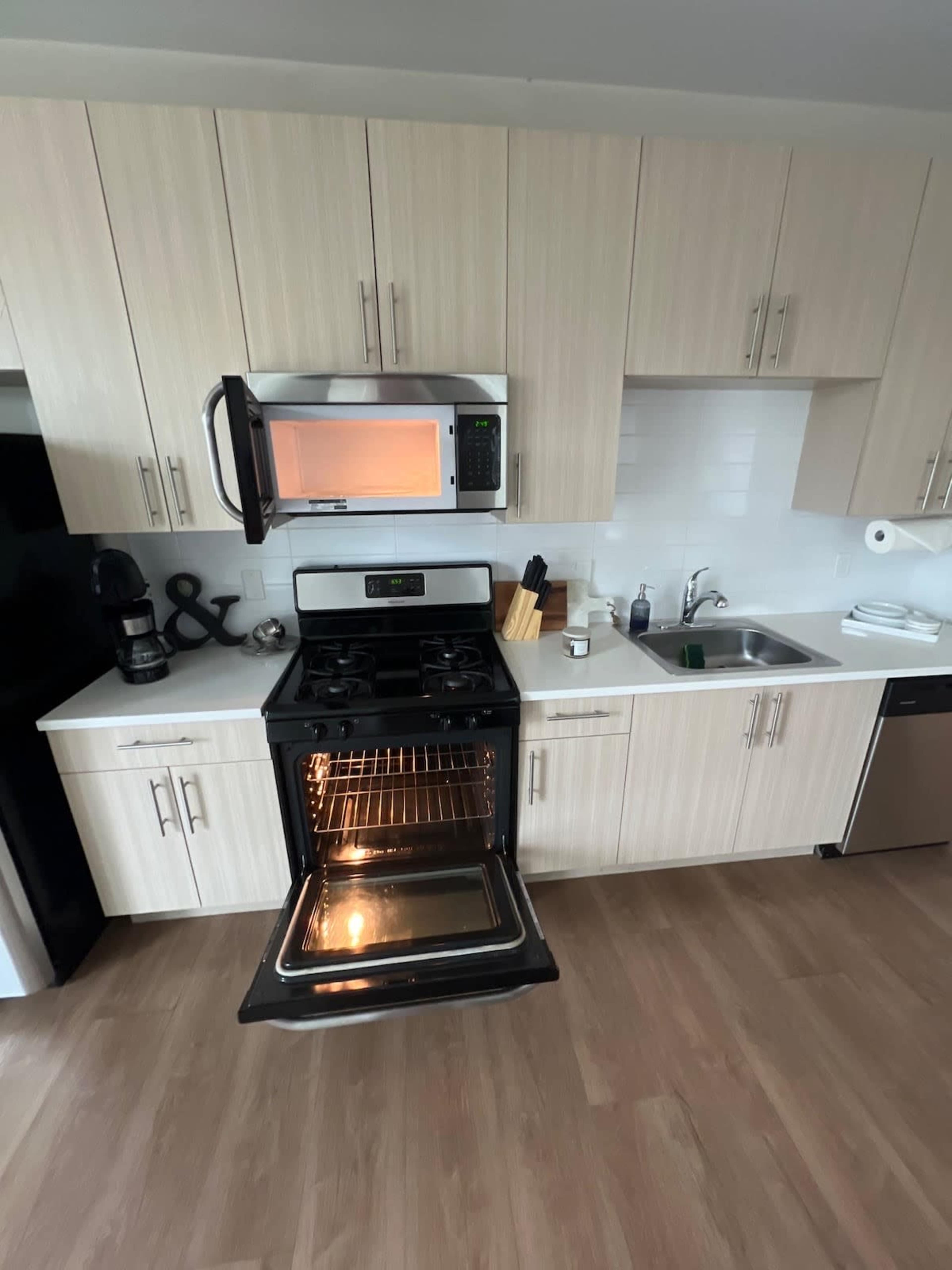 The image shows a modern kitchen with light wood cabinetry, a black oven with its door open, and a microwave above the oven.