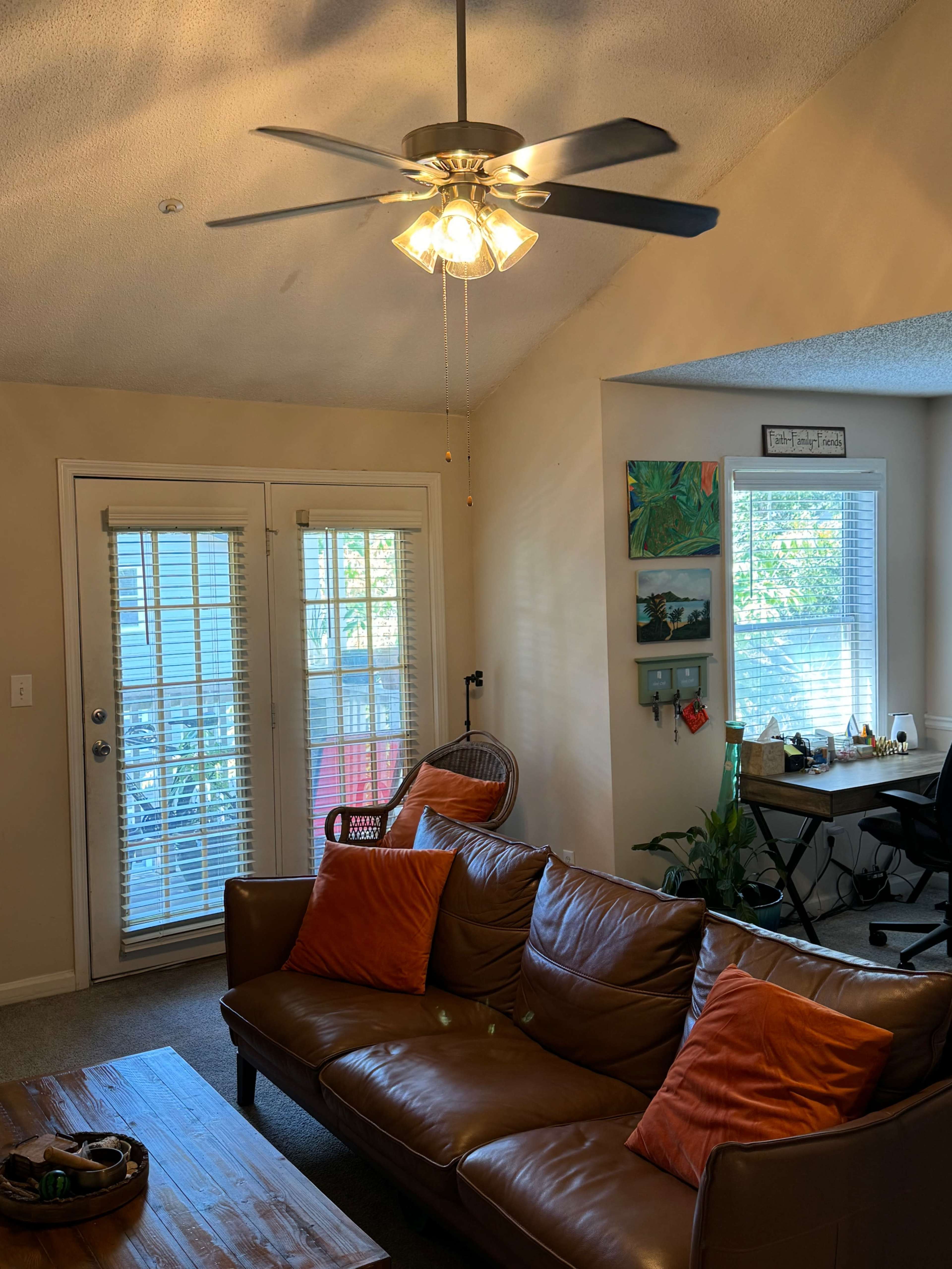 The image shows a cozy living room with a brown leather couch, a ceiling fan, a wooden coffee table, and large windows with blinds.