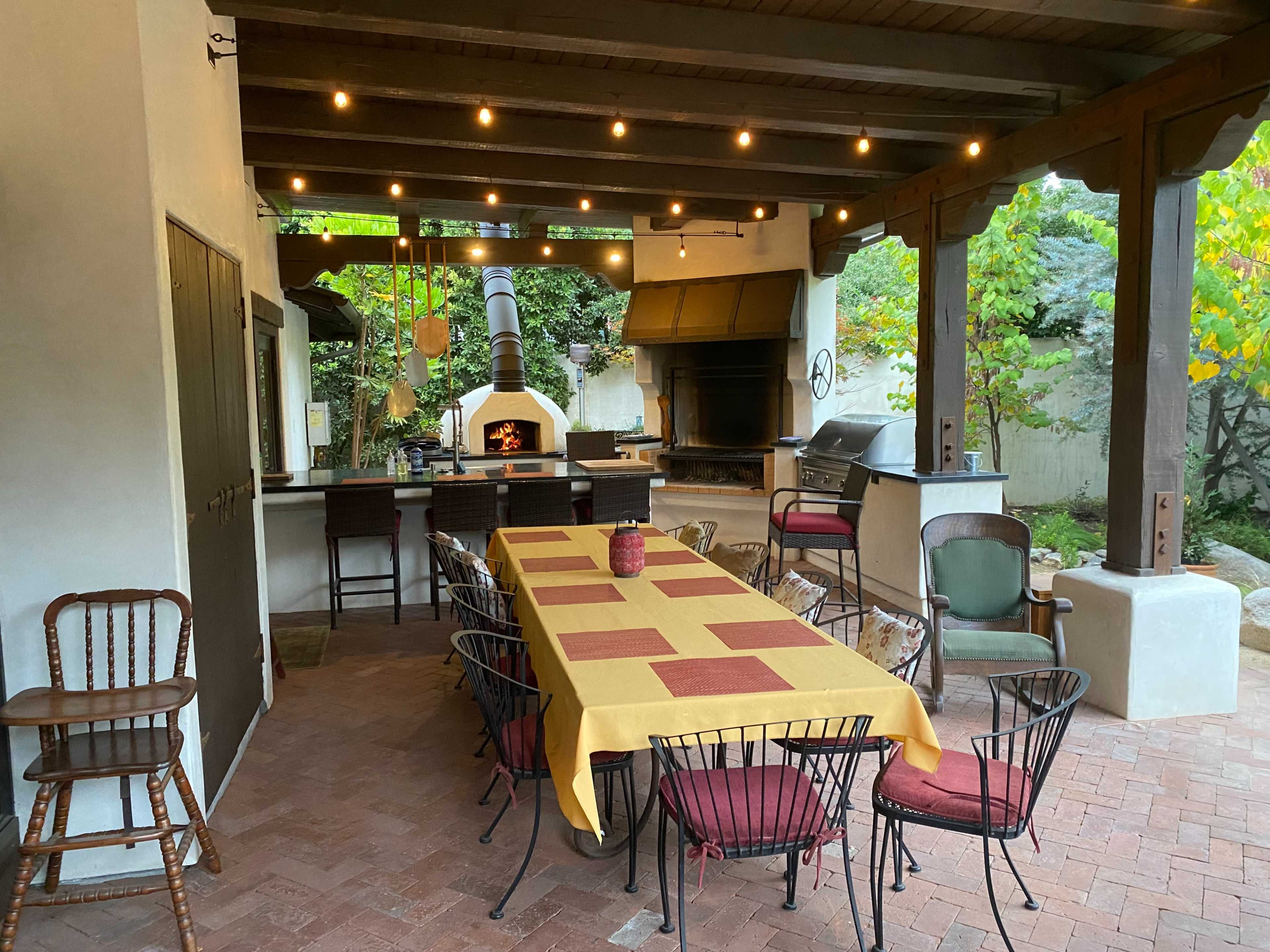 An outdoor dining area features a long table set with a yellow tablecloth, surrounded by chairs, an outdoor oven, and a grill under wooden beams with string lights above.