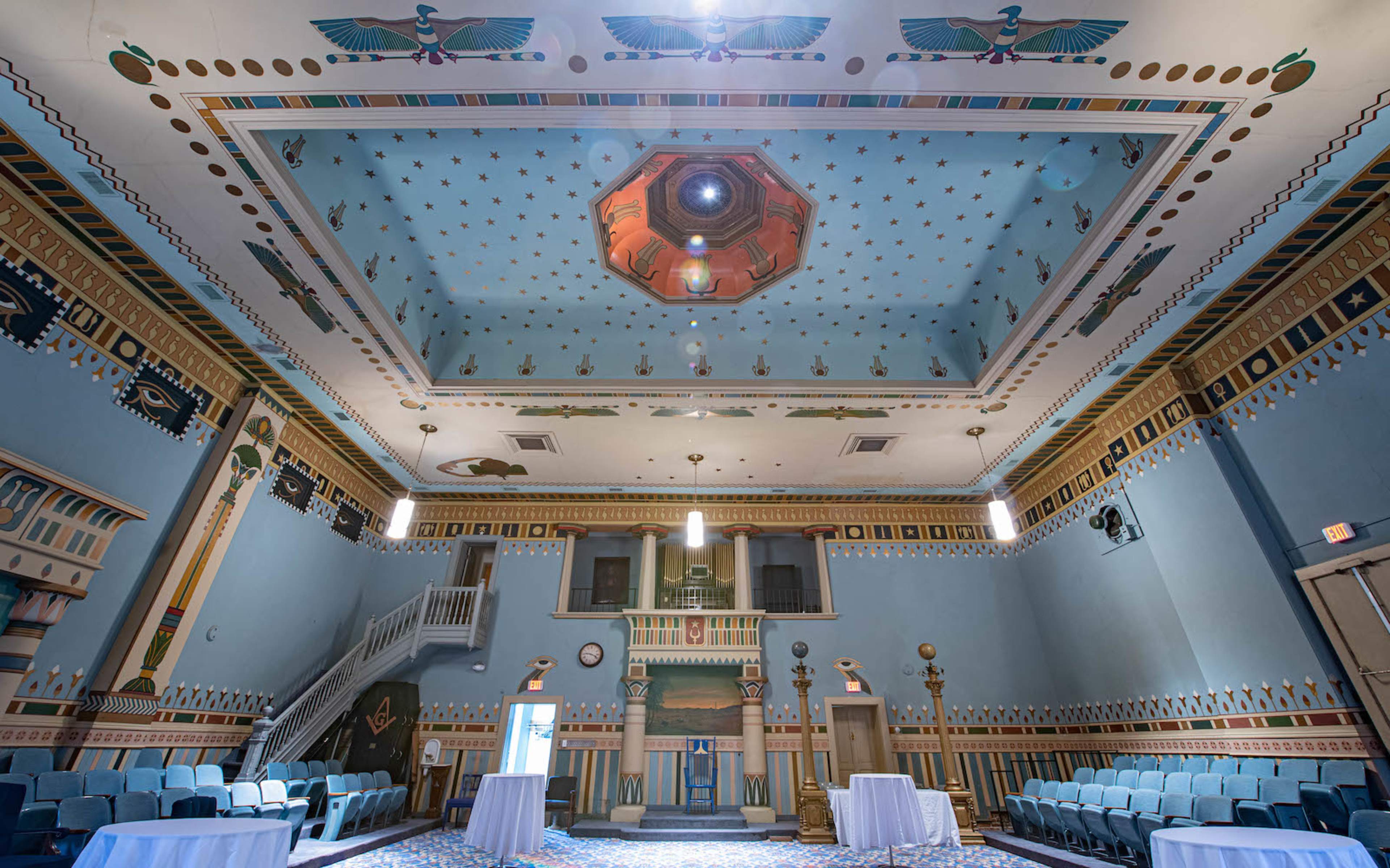 A spacious hall with a decorated ceiling featuring geometric patterns, flanking columns, and tables set up in the center.