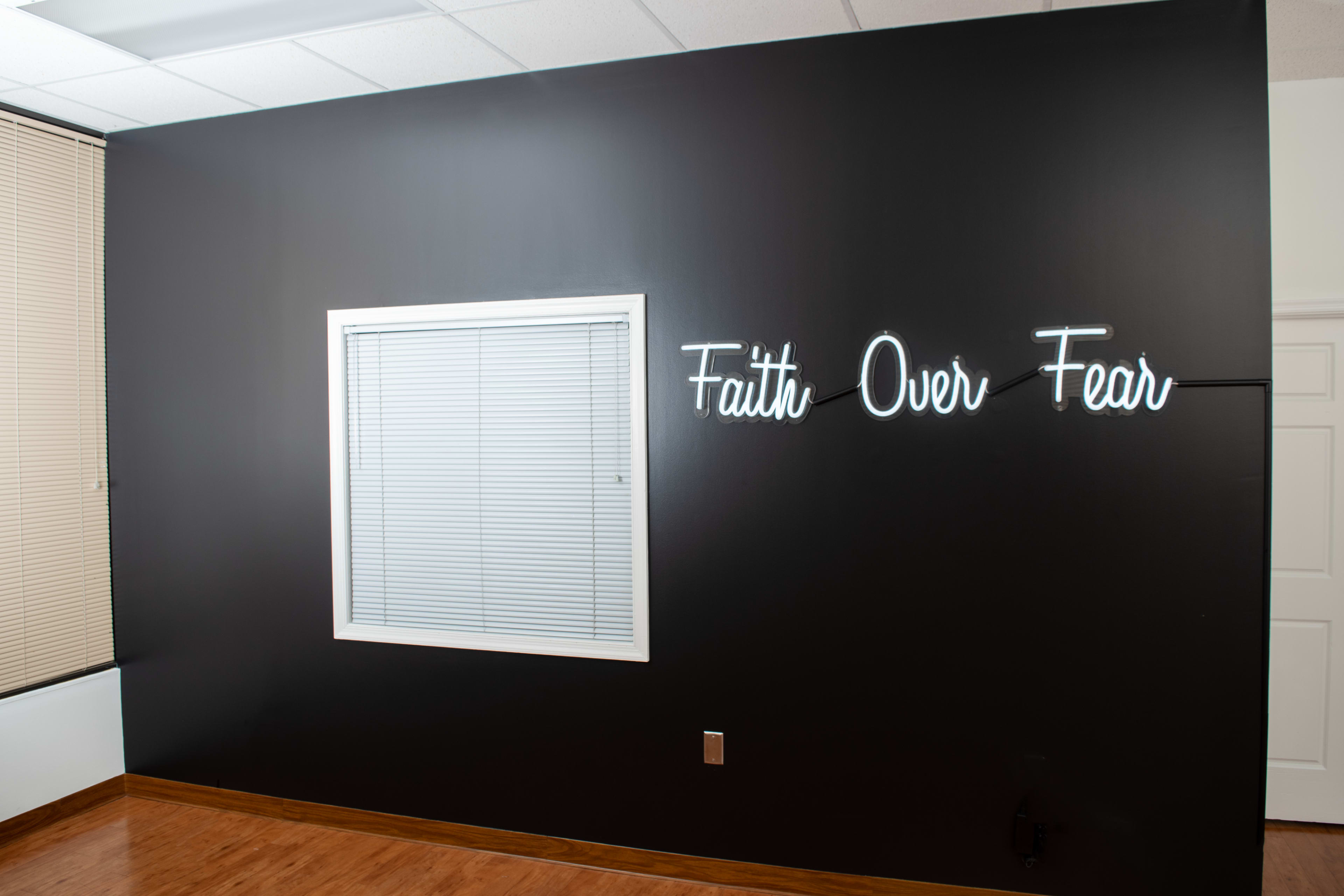 A black wall features a neon sign that reads "Faith Over Fear" next to a window with blinds.