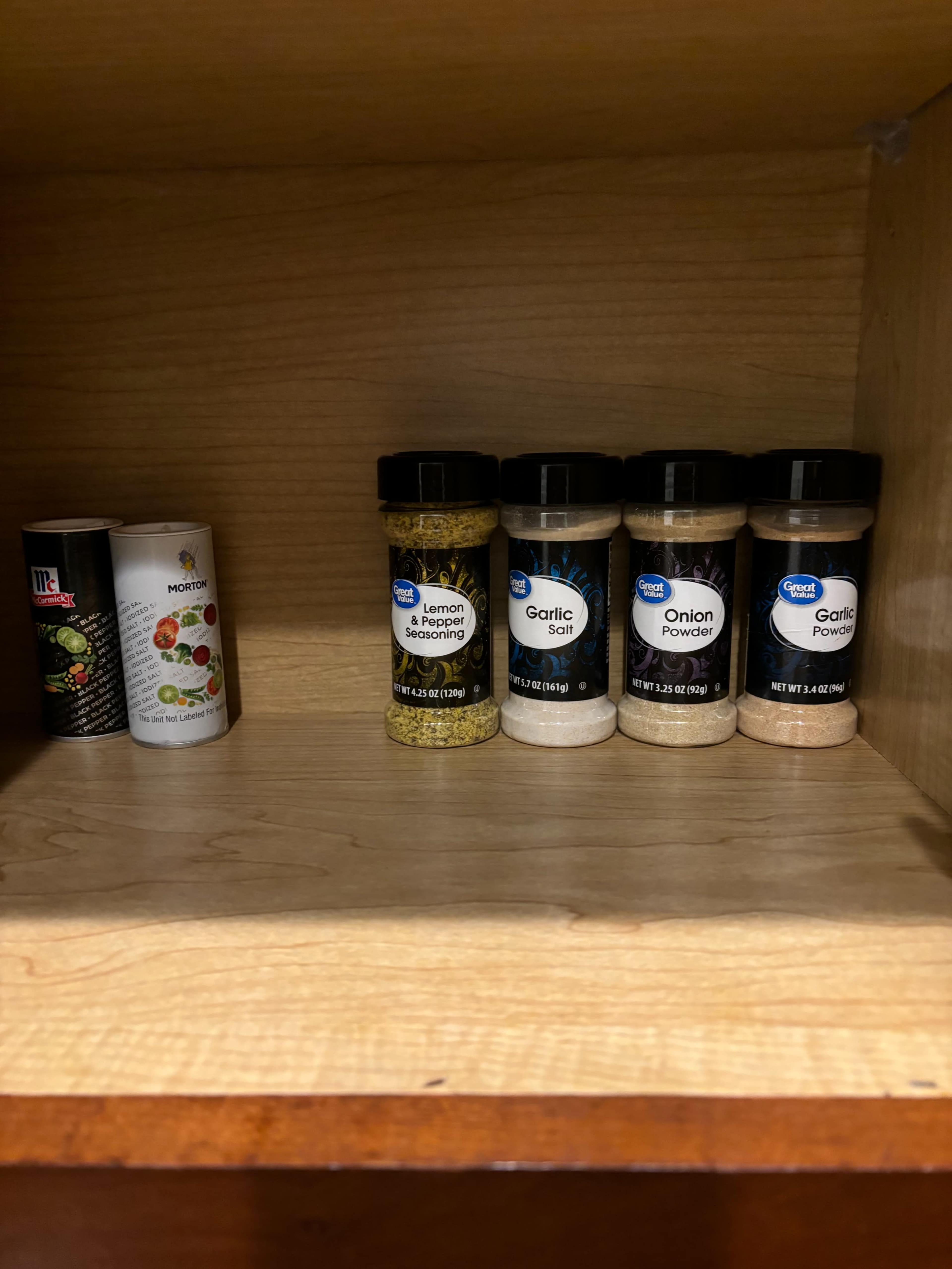 The image shows a wooden cabinet shelf containing six spice containers, including a container of seasoning blend and various powders such as garlic and onion.