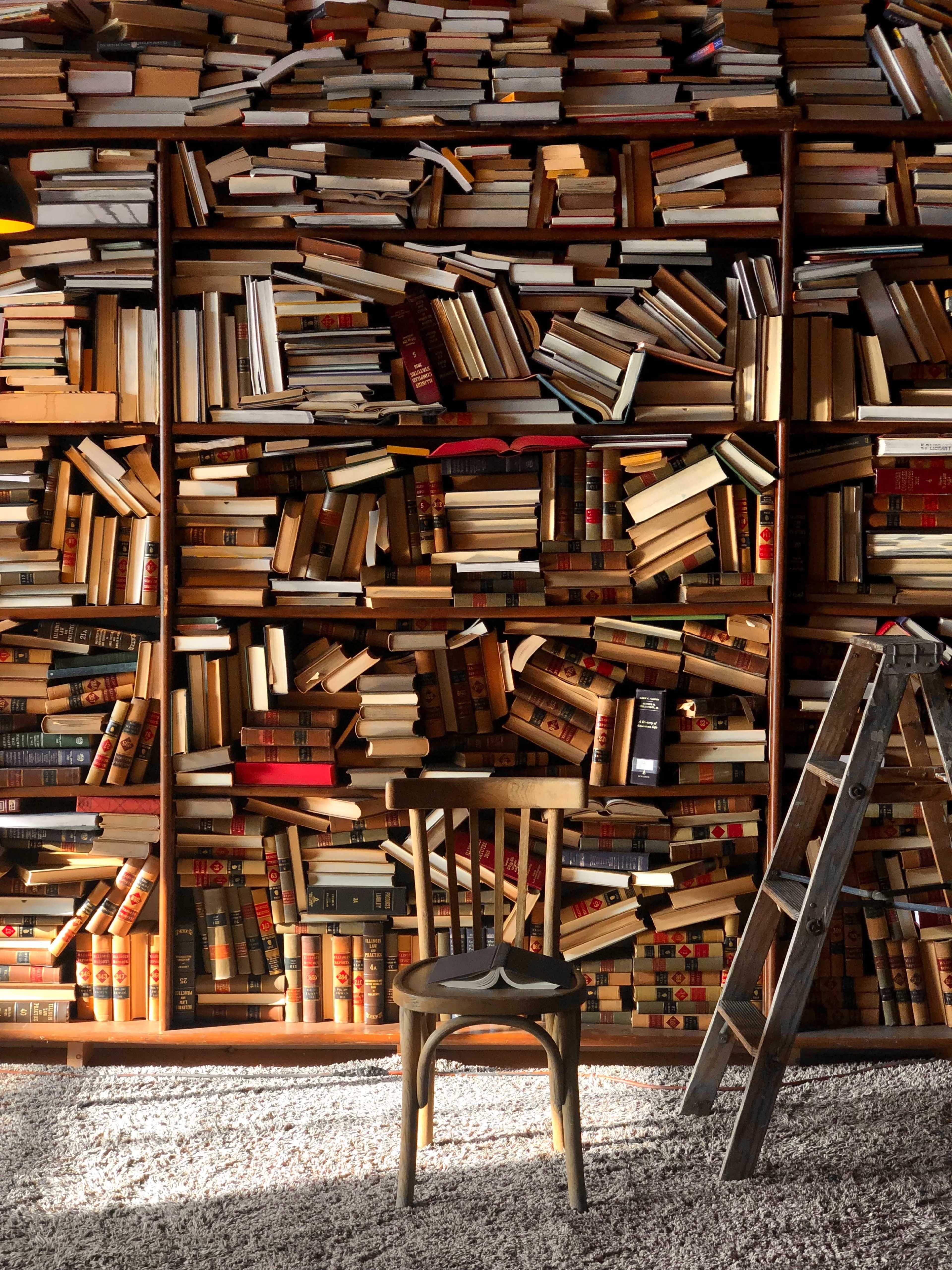 A wooden chair faces a towering bookshelf filled with stacks of books in a well-lit room.