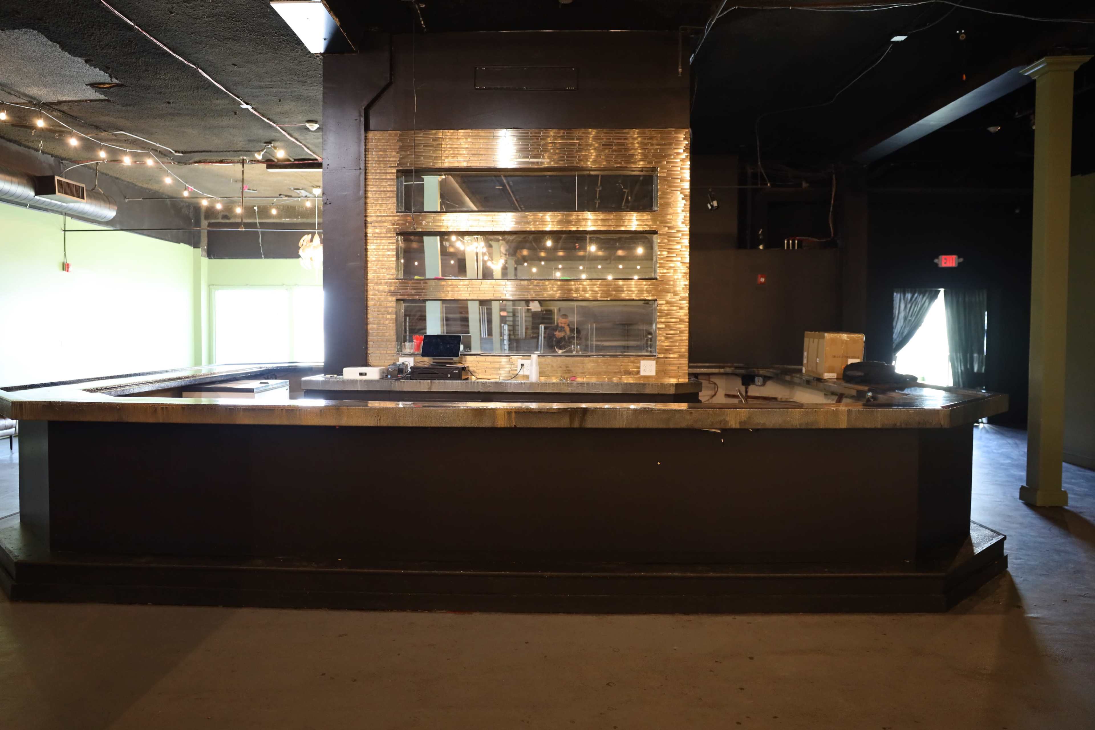 An empty bar with a polished countertop and a large mirror backdrop is shown in a dimly lit venue.