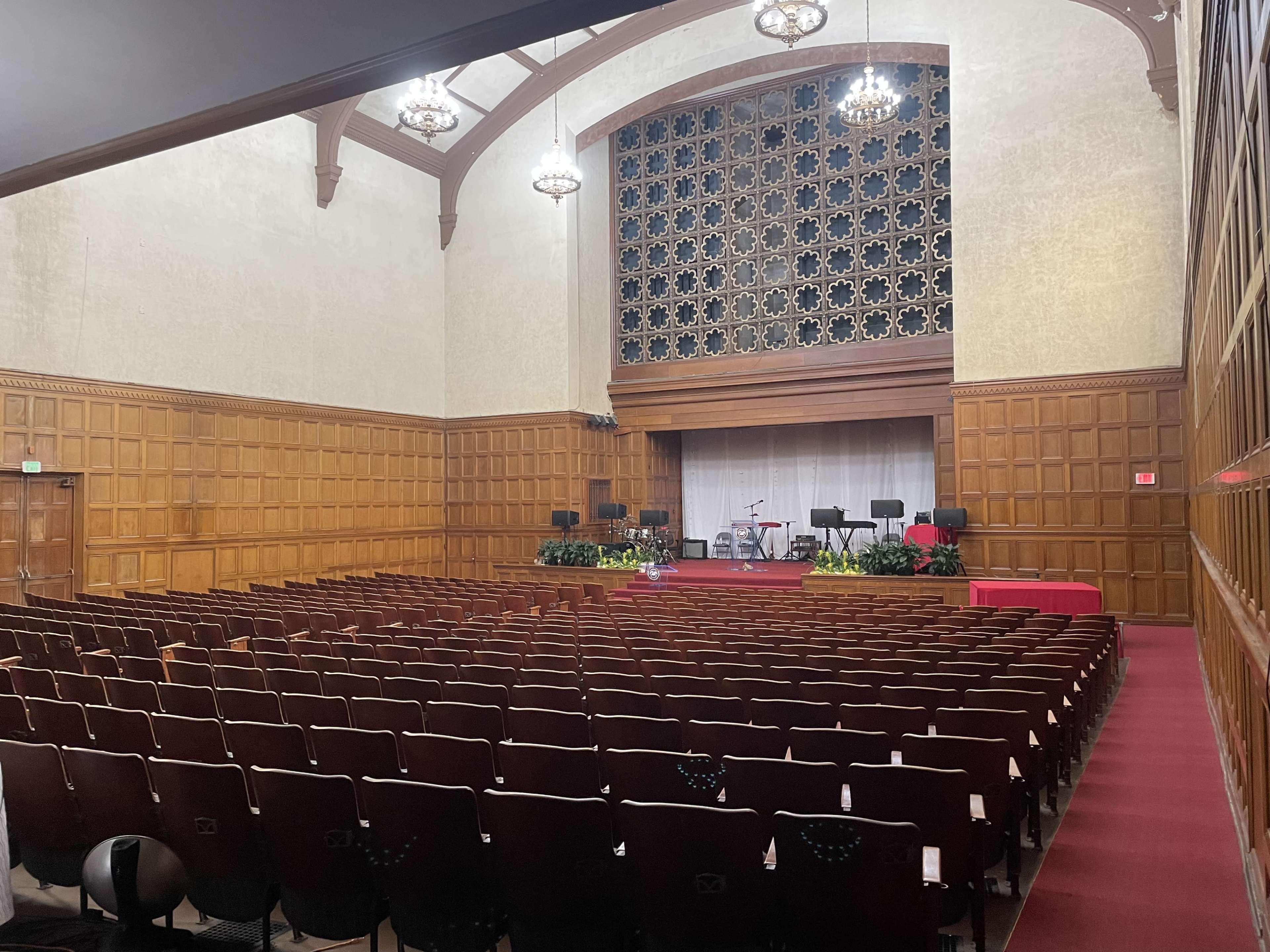 The image shows an empty auditorium with wooden walls, arranged seating, and a stage at the front.