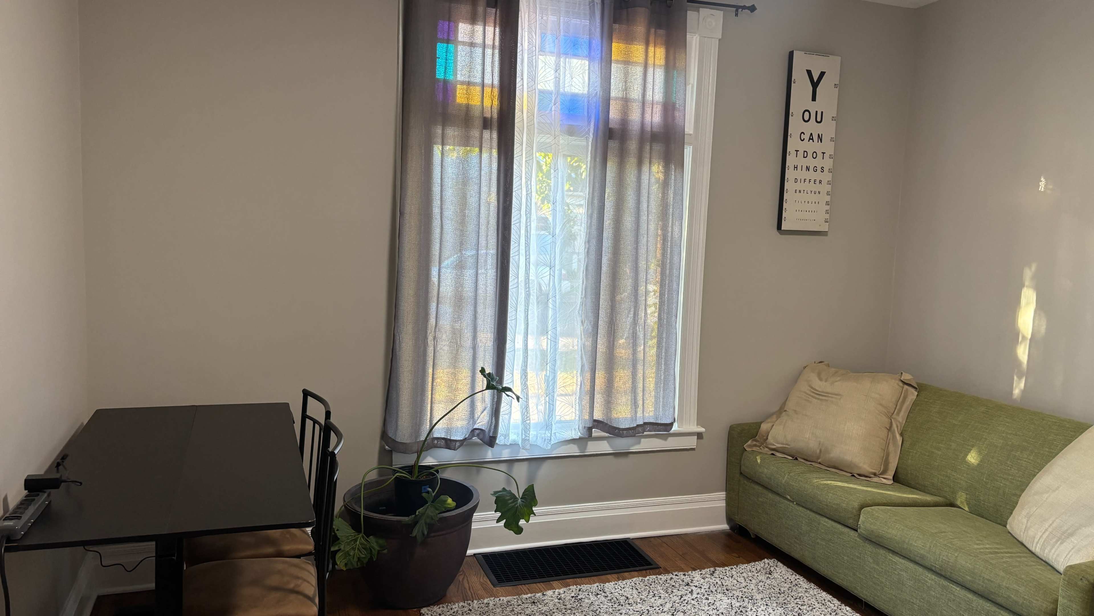 The image shows a living room with a green couch, a small dining table, and a window covered with sheer curtains that have colorful panels.