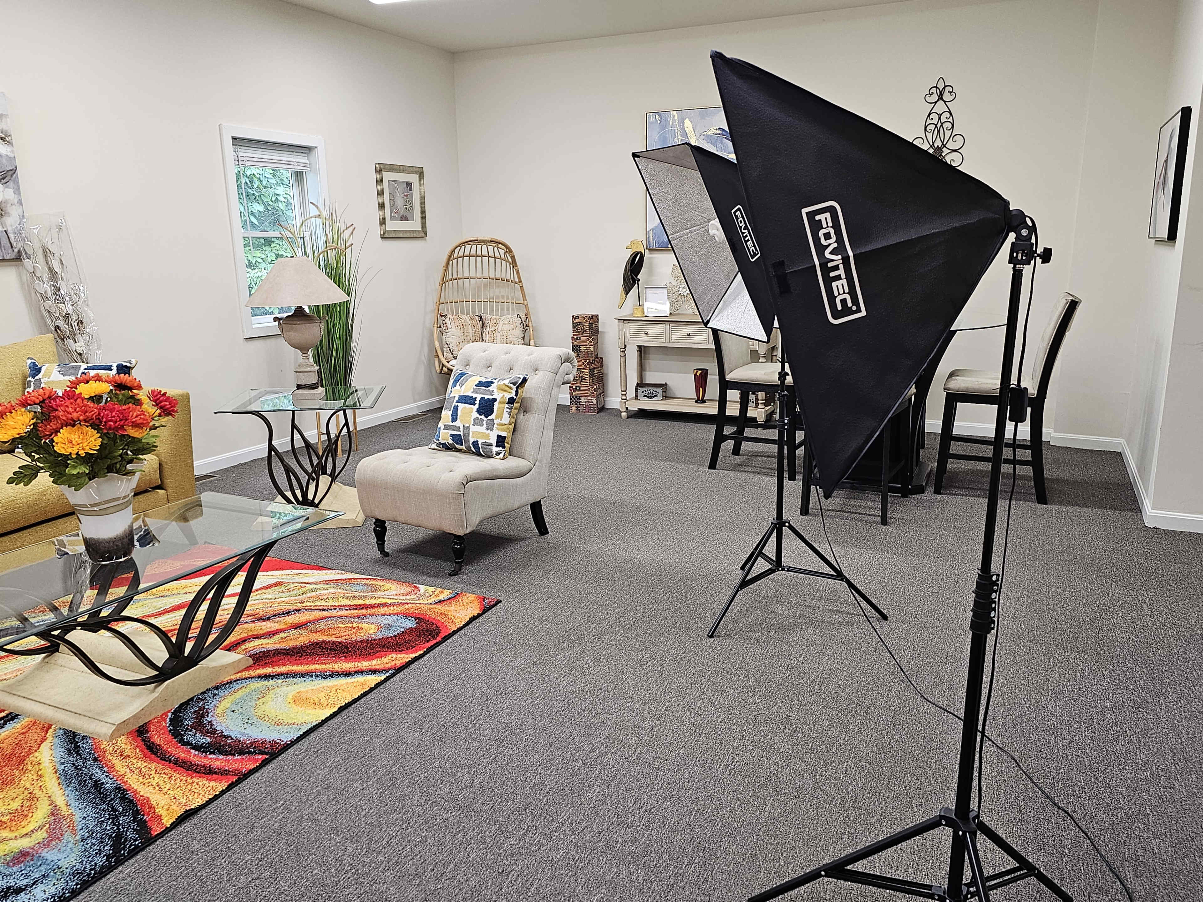 A brightly lit room features a seating area with a yellow sofa, a glass coffee table on a colorful rug, and a corner with a lamp and artwork.