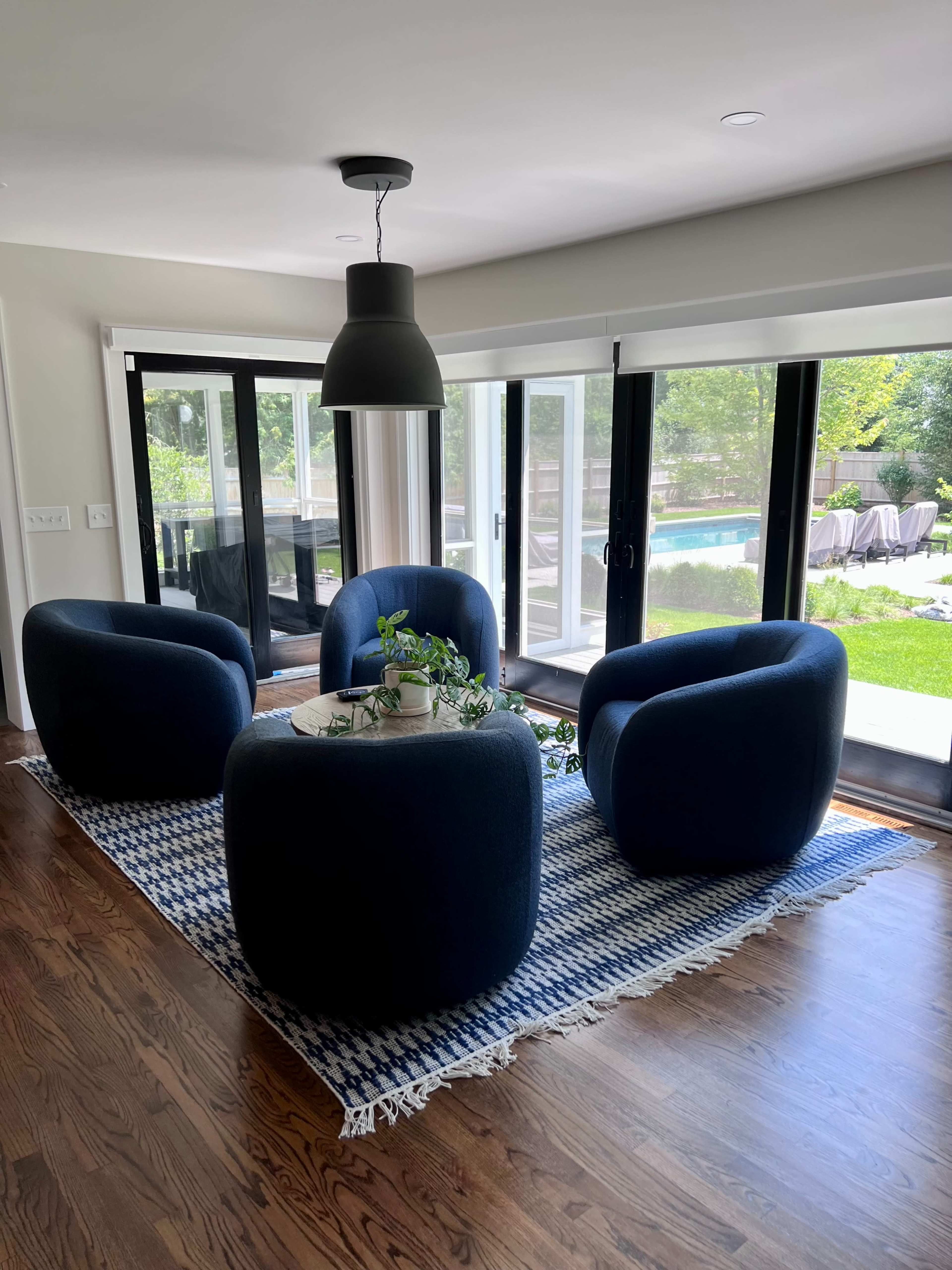 A cozy seating area features three blue, rounded chairs arranged around a small table on a patterned rug, with large windows overlooking a green outdoor space.
